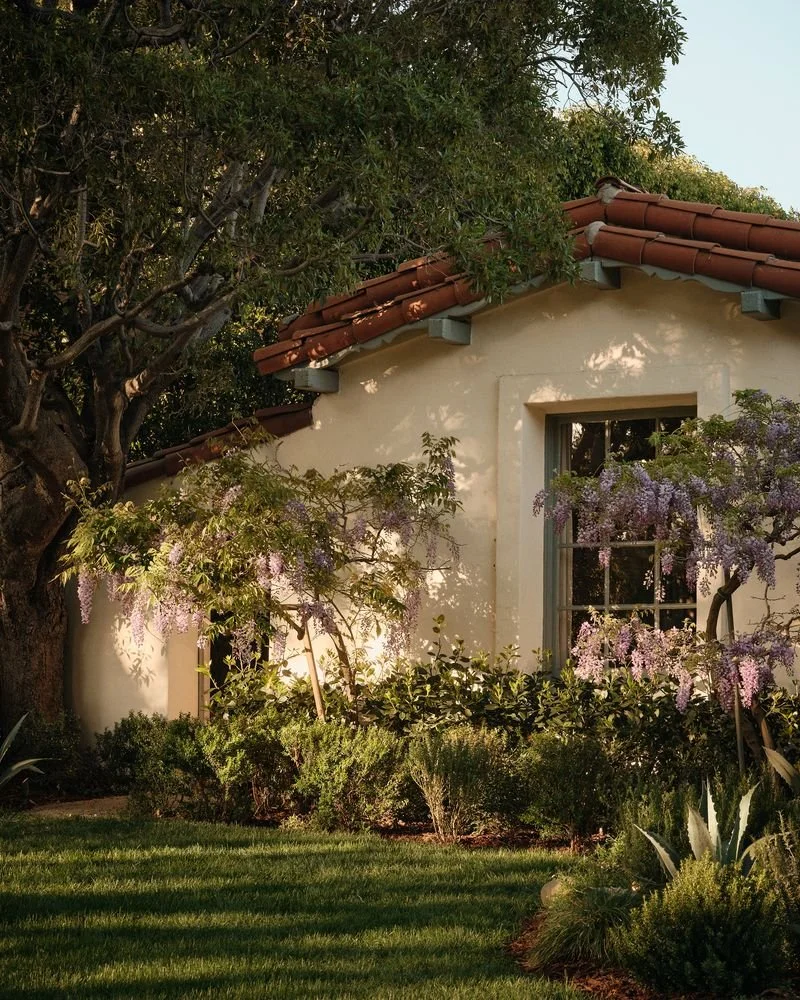 A house with a beige stucco wall, a window, and a terracotta-tiled roof, surrounded by lush greenery and flowering trees in the garden.