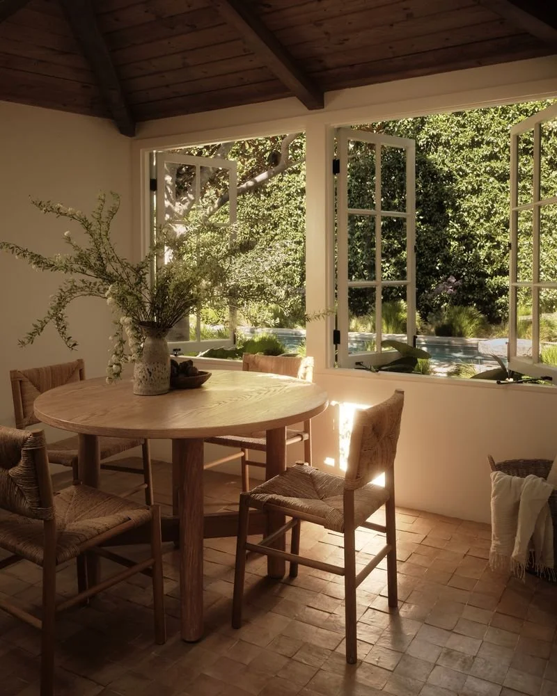 Sunlit dining room with a round wooden table, four woven chairs, a large floral arrangement in a vase, open windows showing greenery and trees outside.