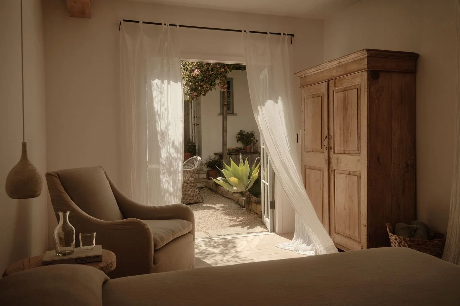A cozy room with an armchair near a small side table, a hanging lamp, and sheer curtains opening to a patio with potted plants and a large agave plant outside.