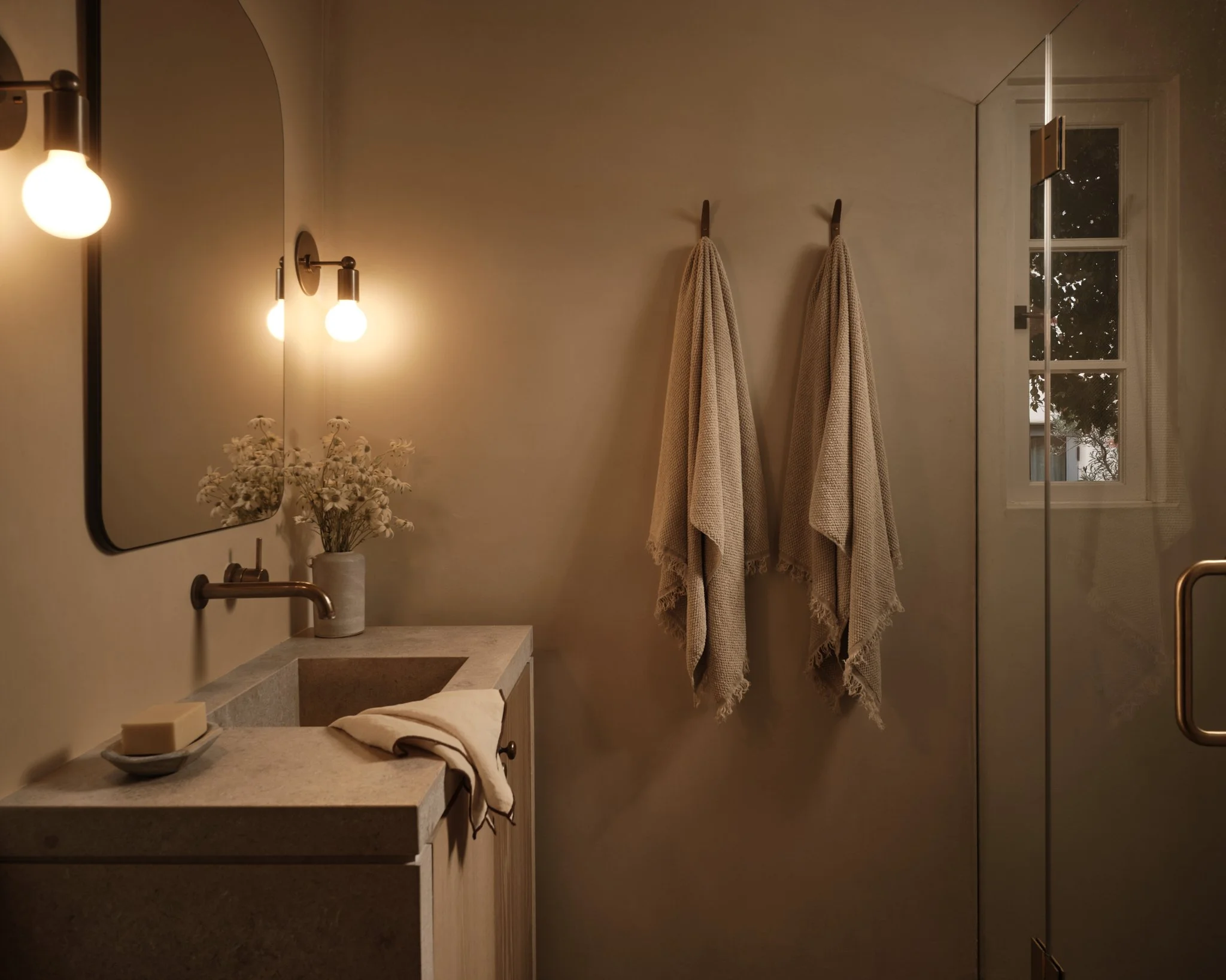 A cozy bathroom with a beige color scheme. There are two wall-mounted light fixtures, a mirror, a small vanity with a beige countertop, a vase with white flowers, and a soap dish with soap. Two beige towels hang on hooks on the wall near a glass-encl