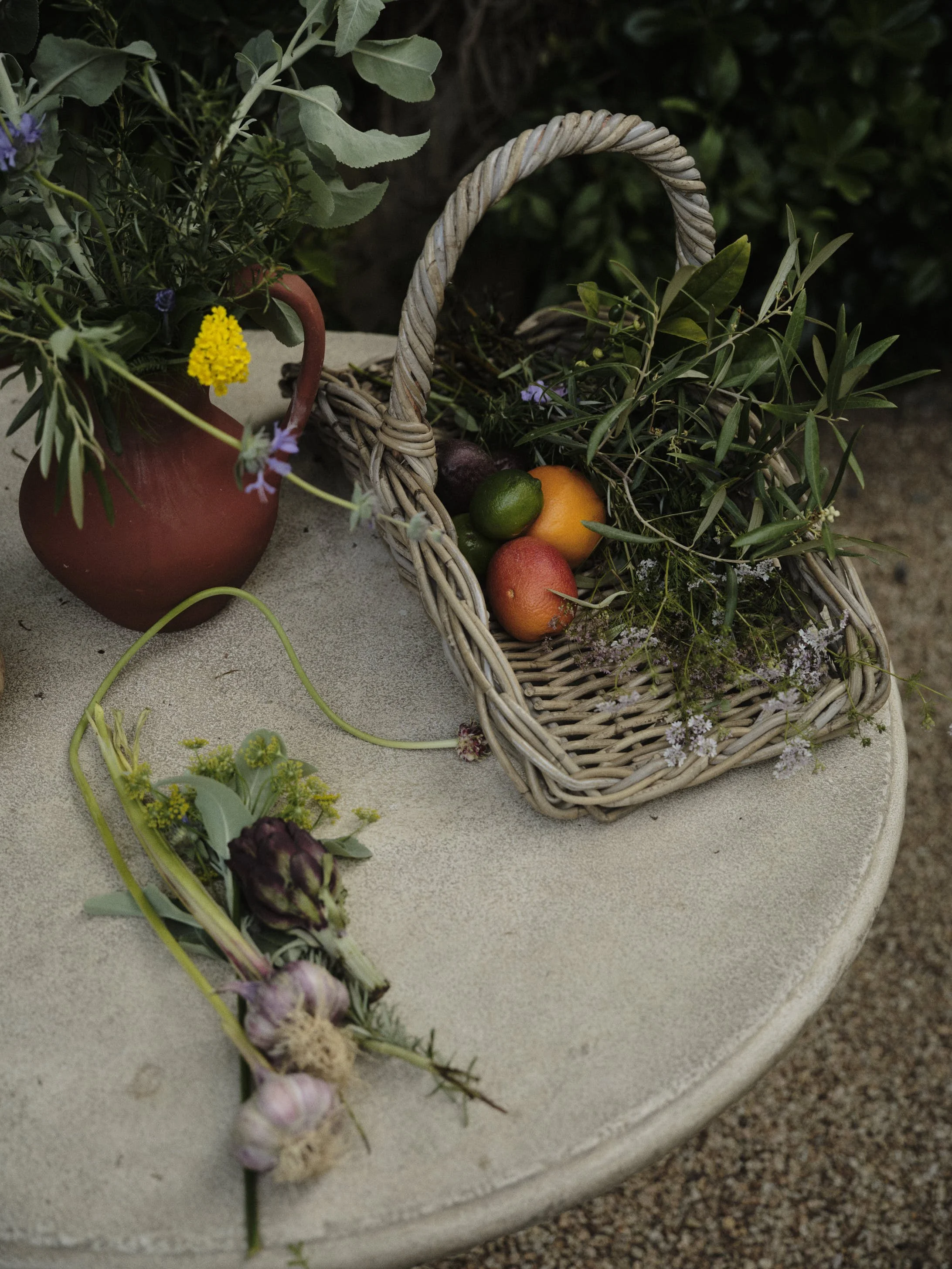 A round table with a small bouquet of flowers in a terracotta vase, and a wicker basket filled with fruits and greenery, outdoor setting.