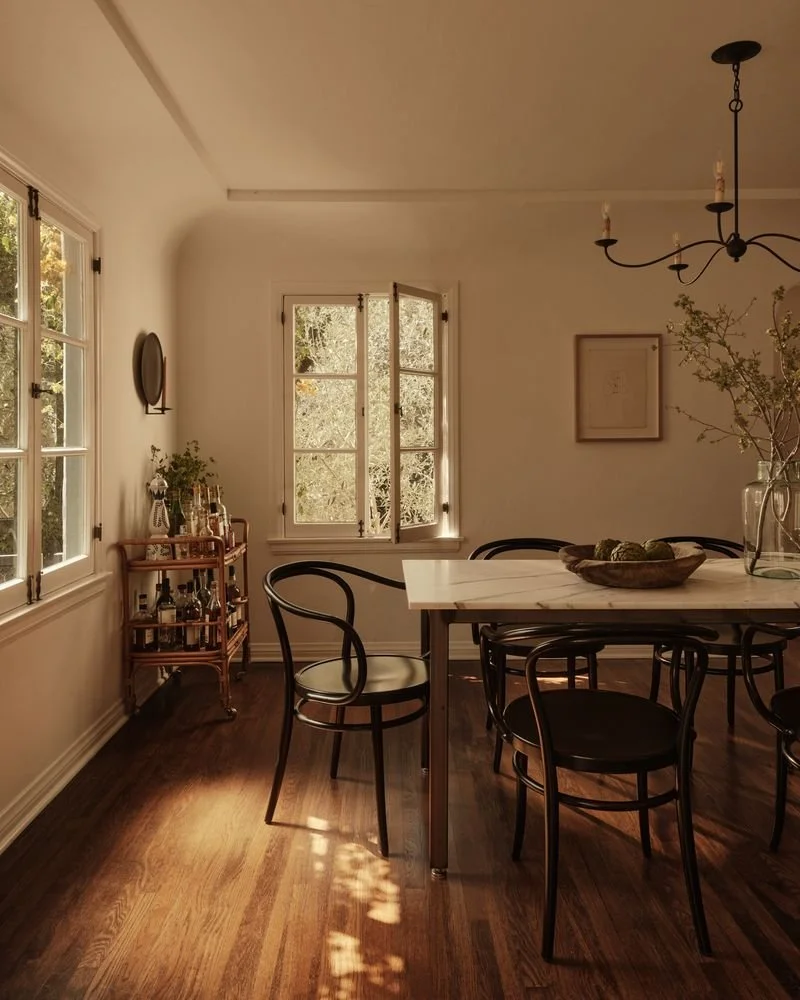 Sunlight streams into a cozy dining room with white walls and wooden floors. There are four black chairs around a white marble table, decorated with a glass vase of branches and a wooden bowl of green vegetables. A small wooden bar cart with bottles 