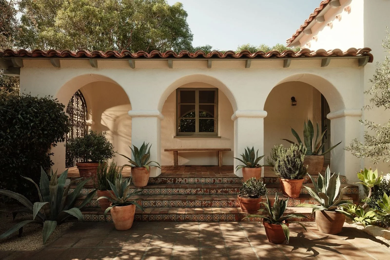 Front porch of a Mediterranean-style house with white stucco walls, terracotta roof tiles, arched entryways, and potted succulents on tiled steps.