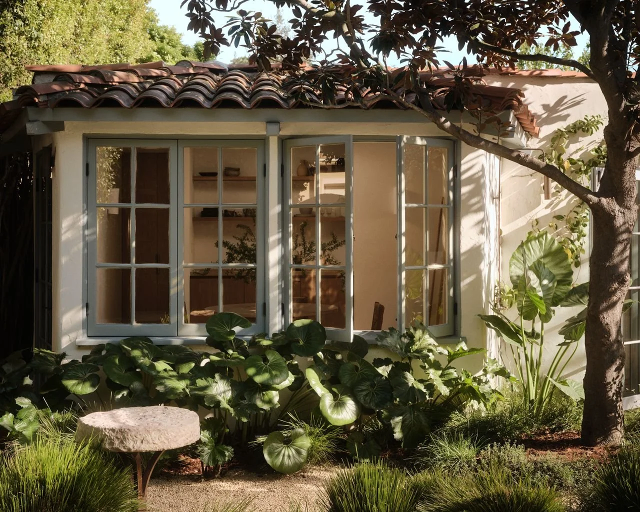 A house with large windowpanes surrounded by lush green plants and a tree in the garden.