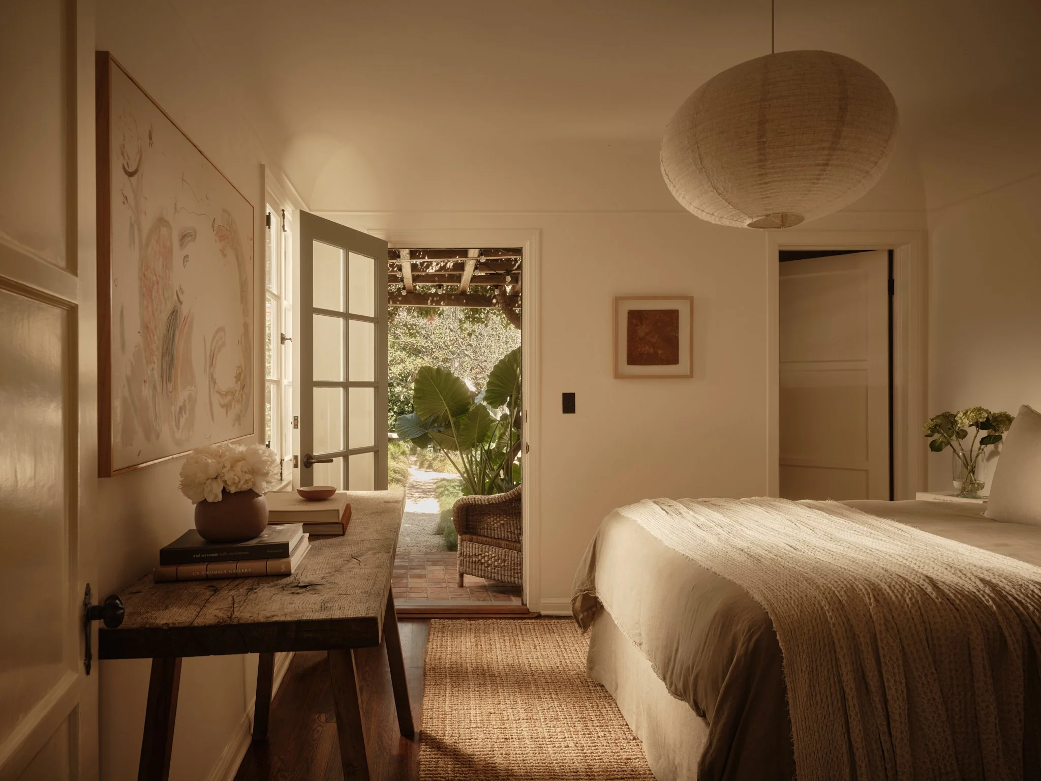 A cozy bedroom with a bed on the right side, a nightstand with a flower vase, and a large paper lantern hanging from the ceiling. An open door leads outside to a garden with greenery and large leaves. A rustic wooden table with books and decor is on 