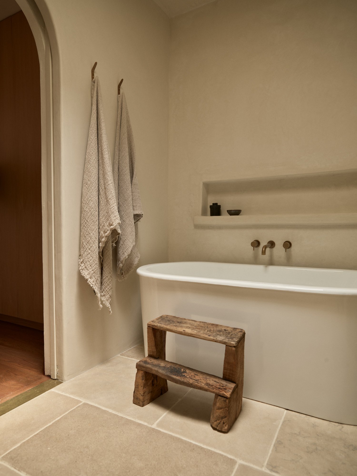 A bathroom with a white bathtub, two beige towels hanging on hooks, a small rustic wooden stool in front of the tub, and a built-in shelf with black and brown containers.