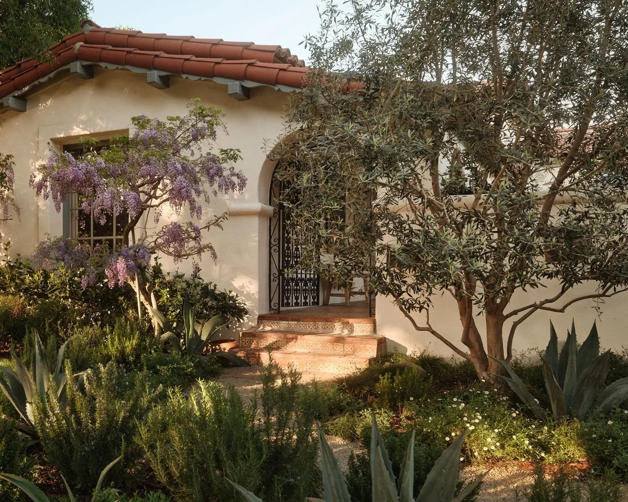 The front of a house with a white stucco exterior, a red tiled roof, and a small staircase leading to a door with a black metal gate. There are various plants and trees, including a flowering purple tree and several succulents along the garden bed.