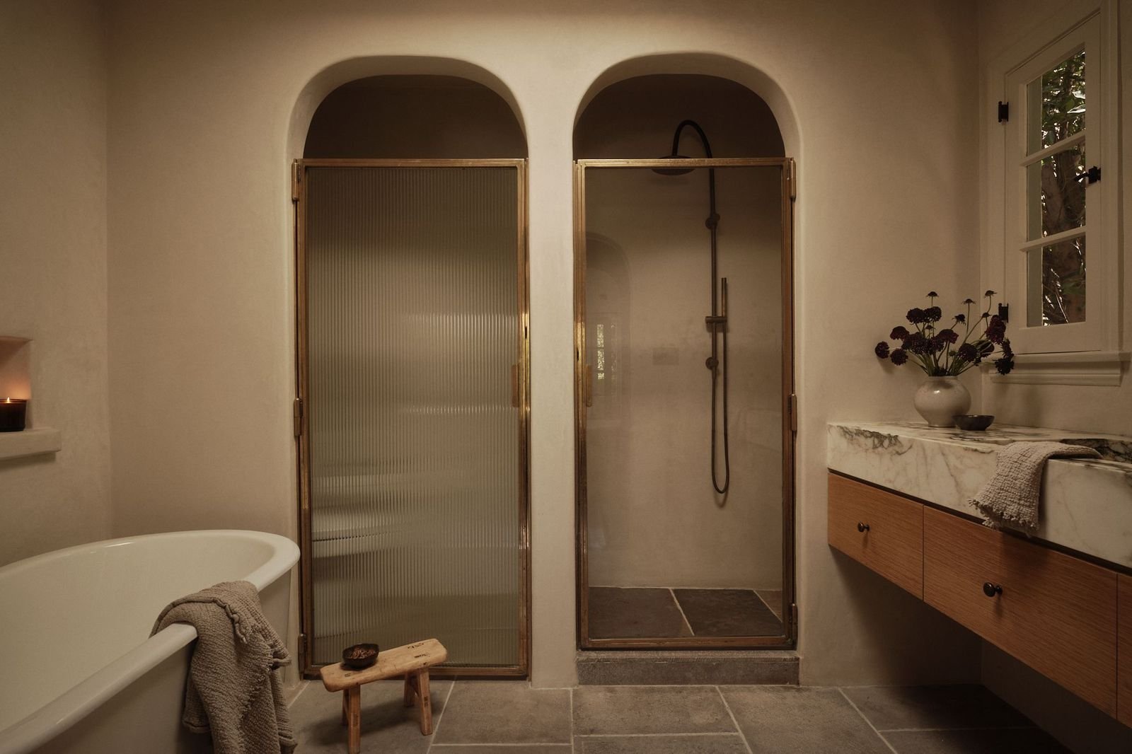 Bathroom with a bathtub on the left, a wooden stool with a soap bar, glass shower doors with brass frames, a shower with black fixtures in the center, a marble countertop with a vase of dark flowers, a towel, and a window with a view of trees outside