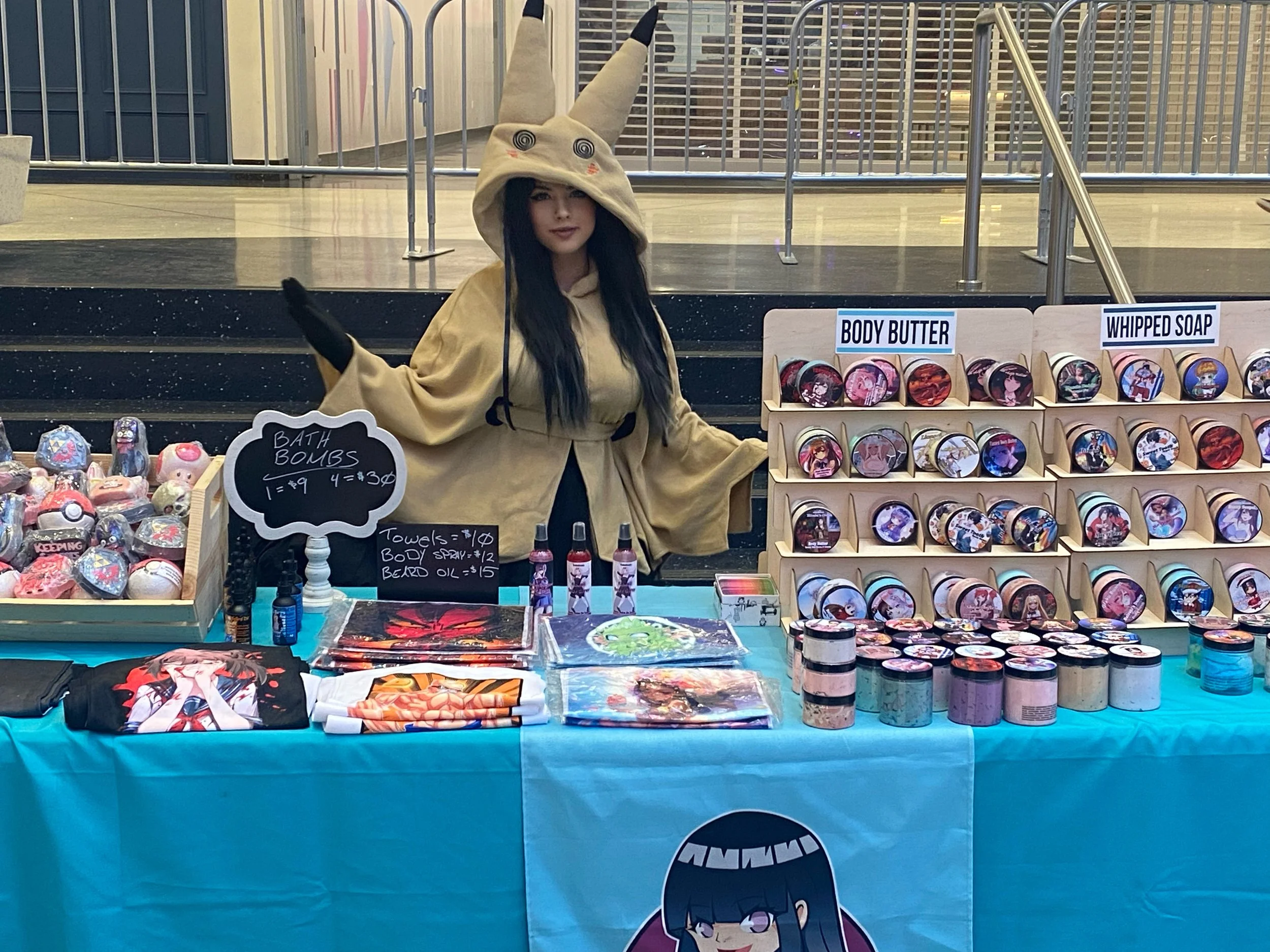 Young woman dressed in a cute costume with a large hat featuring a face with spiral eyes and a small mouth, standing behind a booth selling anime-themed merchandise including body butter, whipped soap, towels, and artwork, with colorful packaging and items on display.