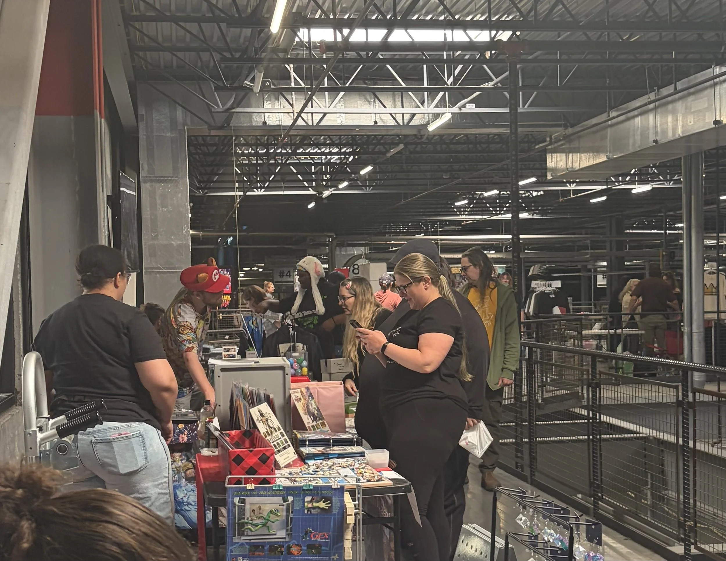 People browsing and shopping at a vendor booth inside a large warehouse or convention center with high ceilings and metal beams.
