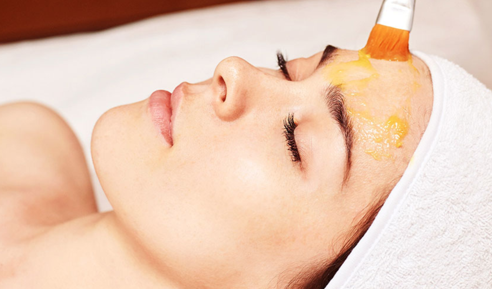 A woman receiving a facial treatment, with a brush applying a yellow facial mask to her forehead, lying down with eyes closed and a white towel wrapped around her head.