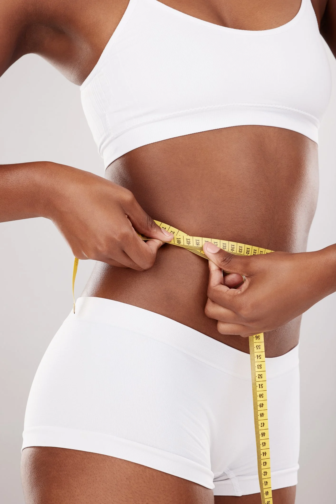 A woman wearing a white sports bra and white shorts measuring her waist with a yellow tape measure.