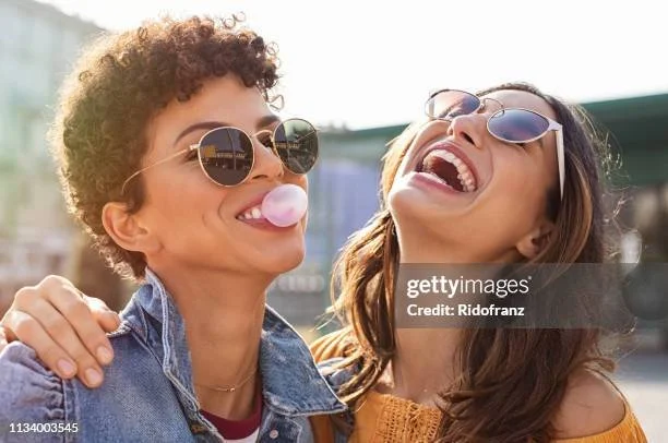 Two women wearing sunglasses and smiling, with one blowing a pink bubble gum bubble, outdoors on a sunny day.