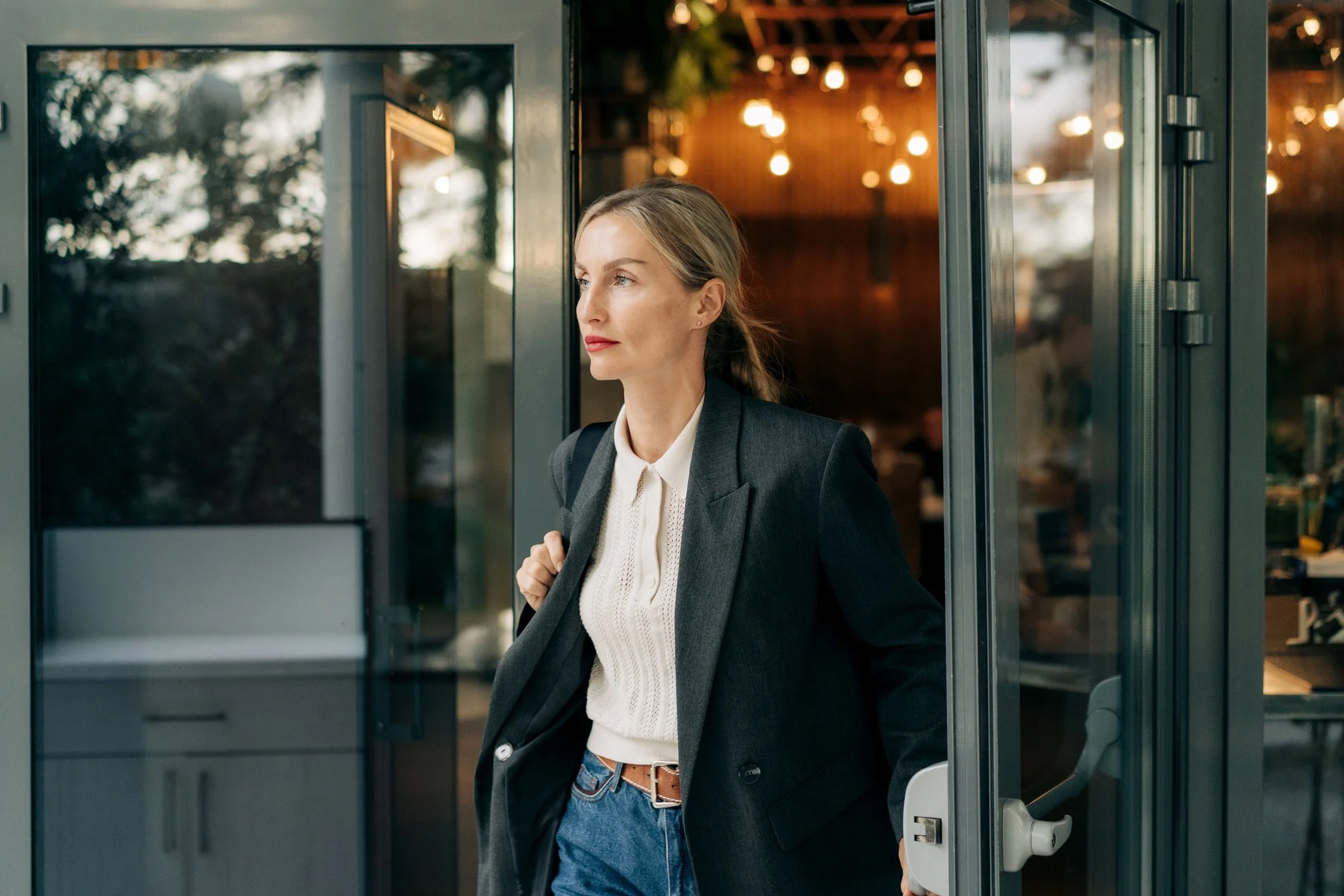A woman with blonde hair, wearing a gray blazer, white blouse, and blue jeans, is standing at the entrance of a building, holding the door open with her right hand. She appears to be in her late 20s or early 30s, with a calm and focused expression, looking to her right. The background shows a warm, illuminated interior with hanging lights.