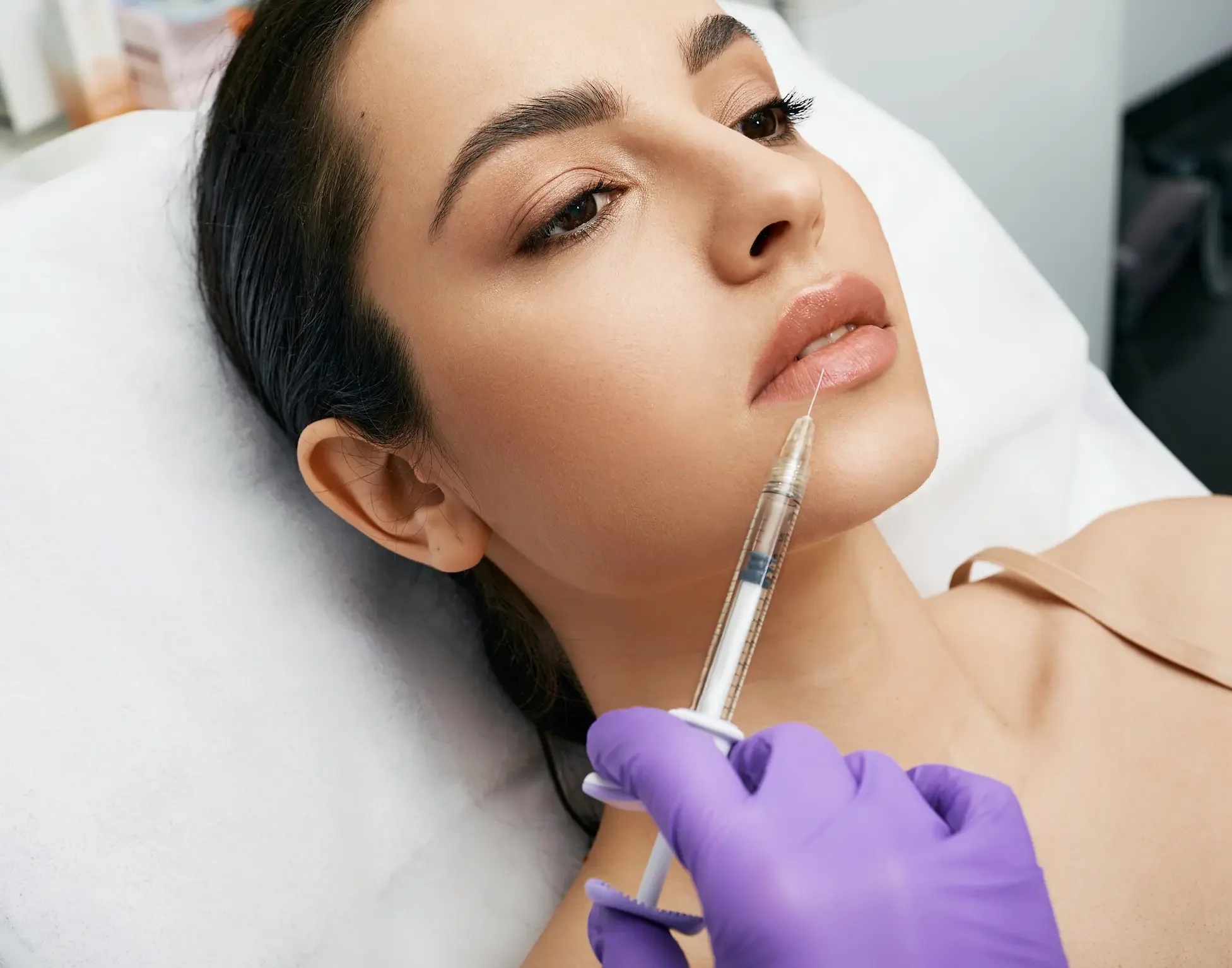 A woman lying on a medical bed receiving a cosmetic injection to her lips from a healthcare professional wearing purple gloves.