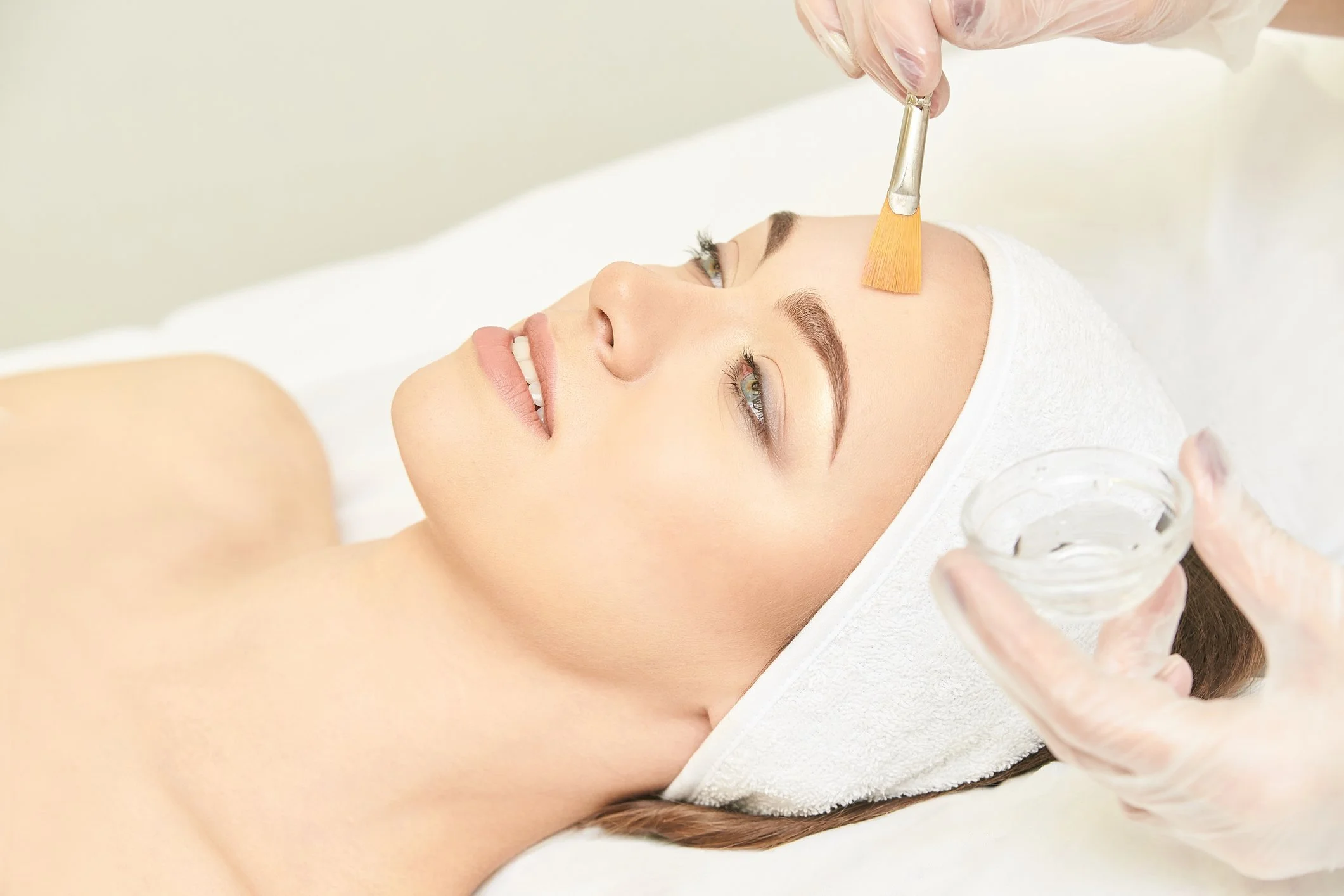 A woman receiving a facial treatment, with a skincare professional applying a facial mask using a brush, while the woman lies on a treatment bed with a towel wrapped around her head.