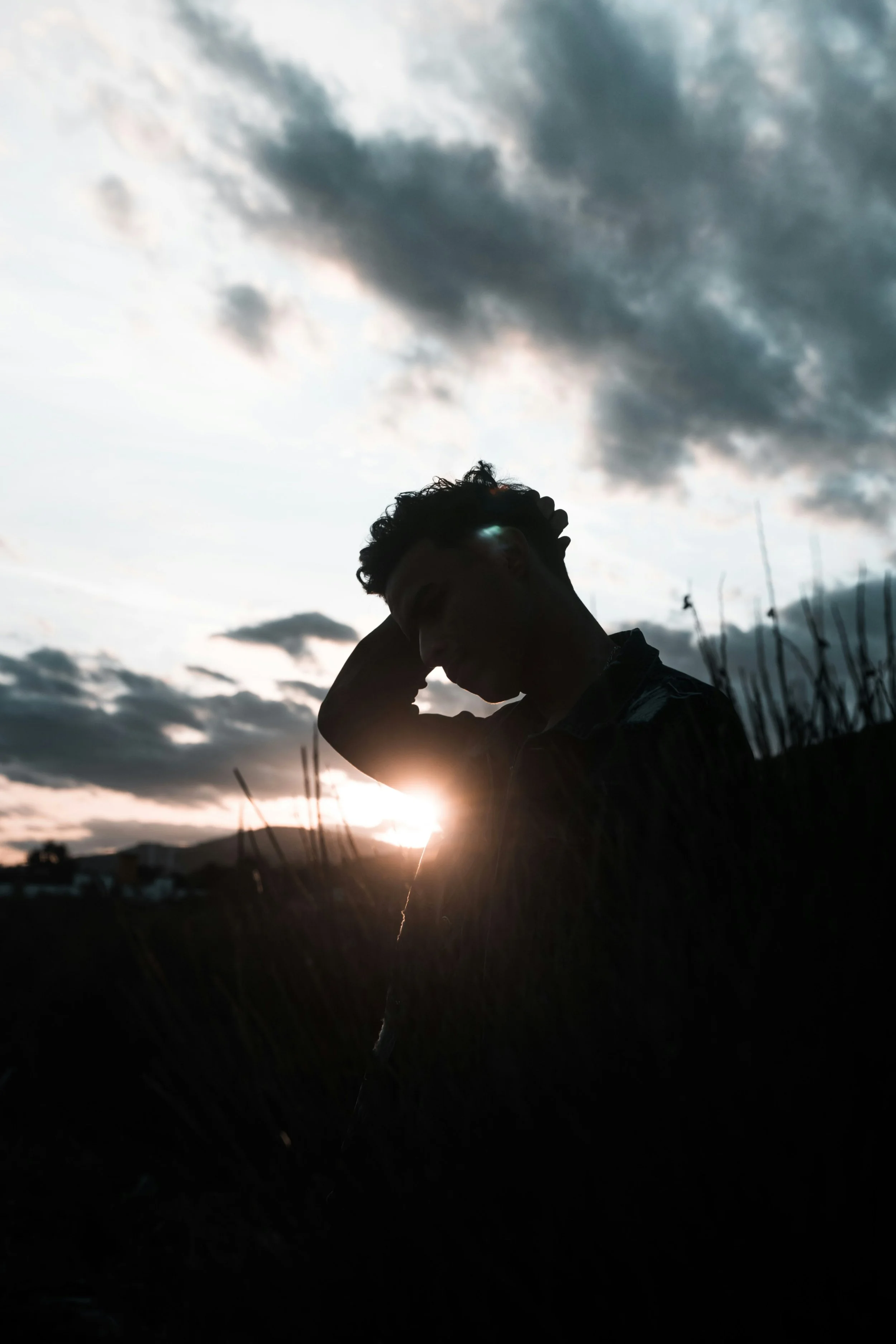 Silhouette of a person with their hand behind their head, standing outdoors during sunset with a cloudy sky in the background.