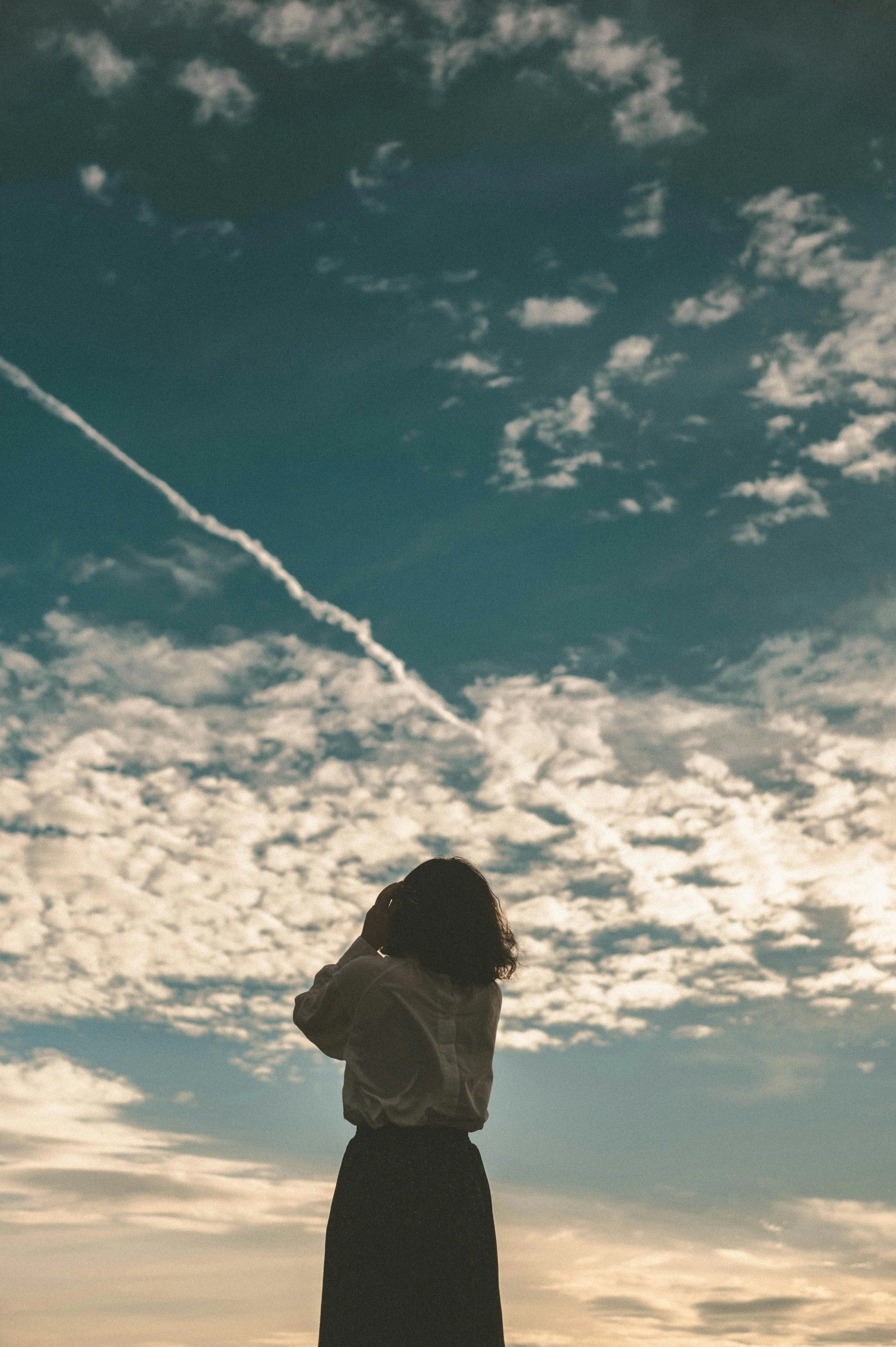 A woman standing outdoors with cloudy sky and contrail in the background.