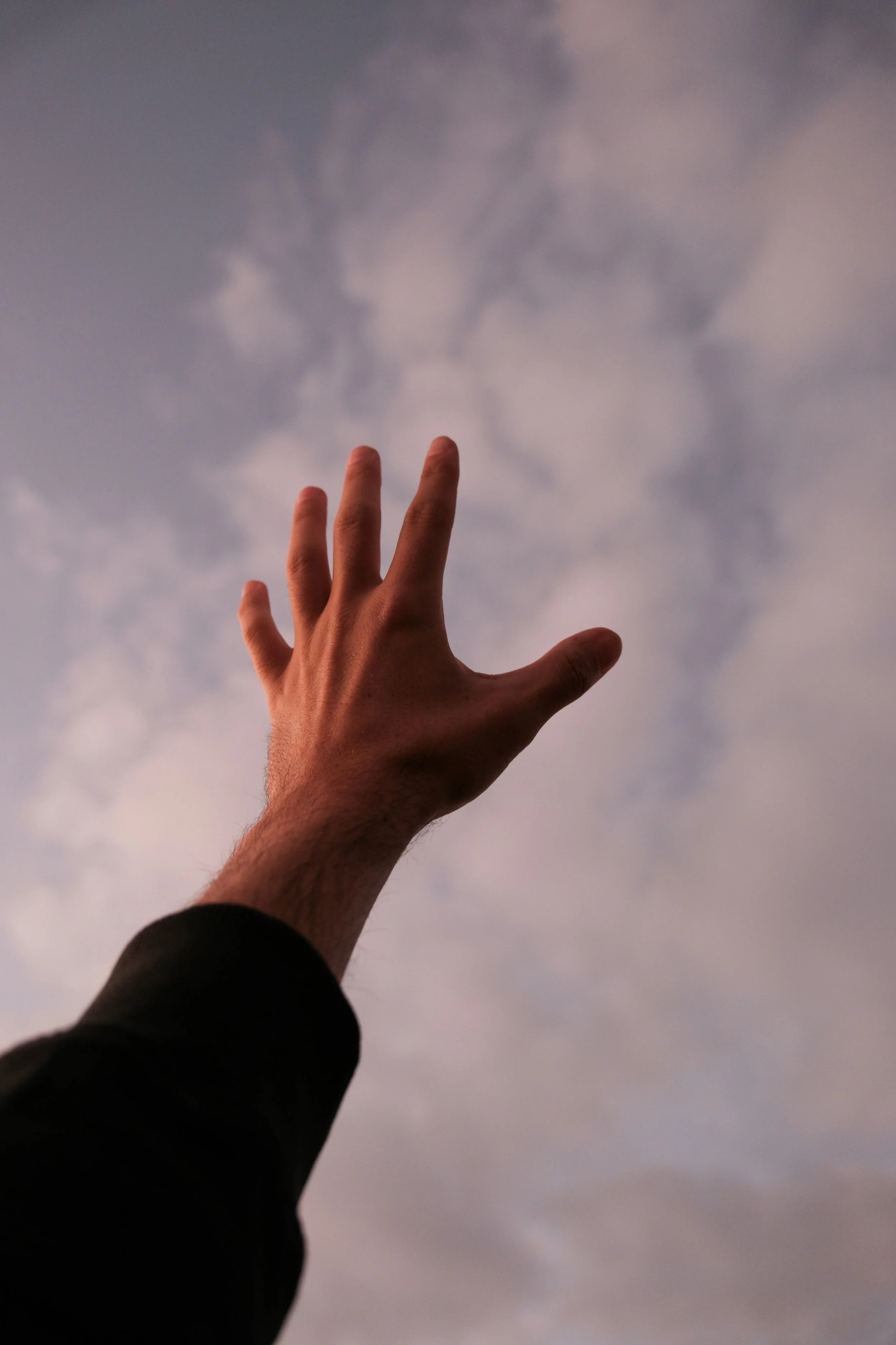A person's hand reaching towards a cloudy sky during sunset or sunrise.