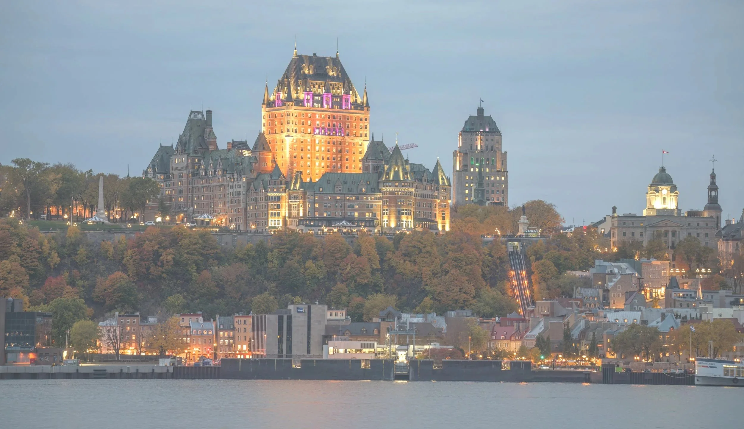 Night view of a castle-like hotel illuminated on a hillside over a waterway, with city buildings below and a cloudy sky above.