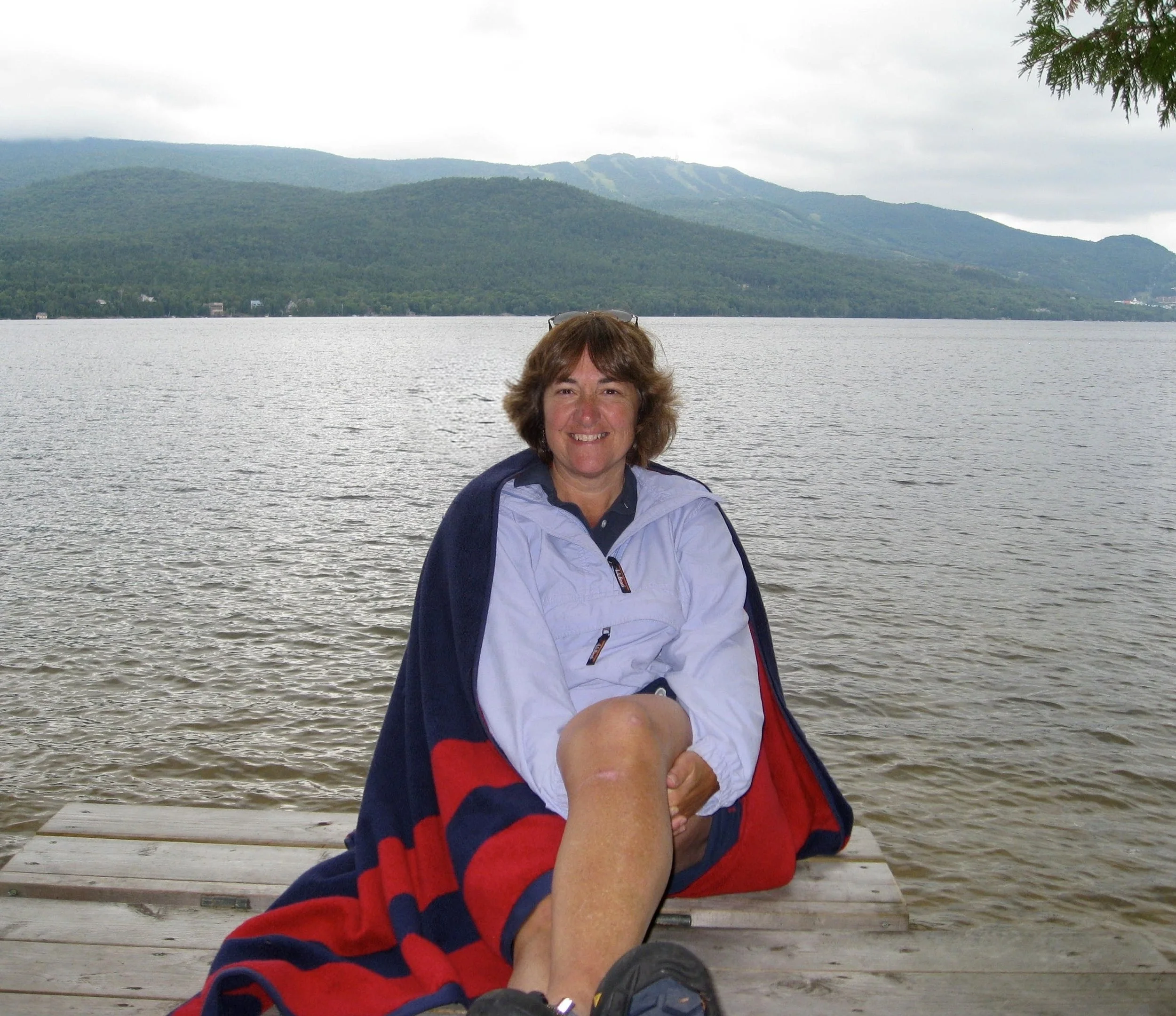 A woman sitting on a wooden dock by a lake, smiling, with a mountain range and cloudy sky in the background. She has brown hair, is wearing a white jacket, and has a blanket around her legs.
