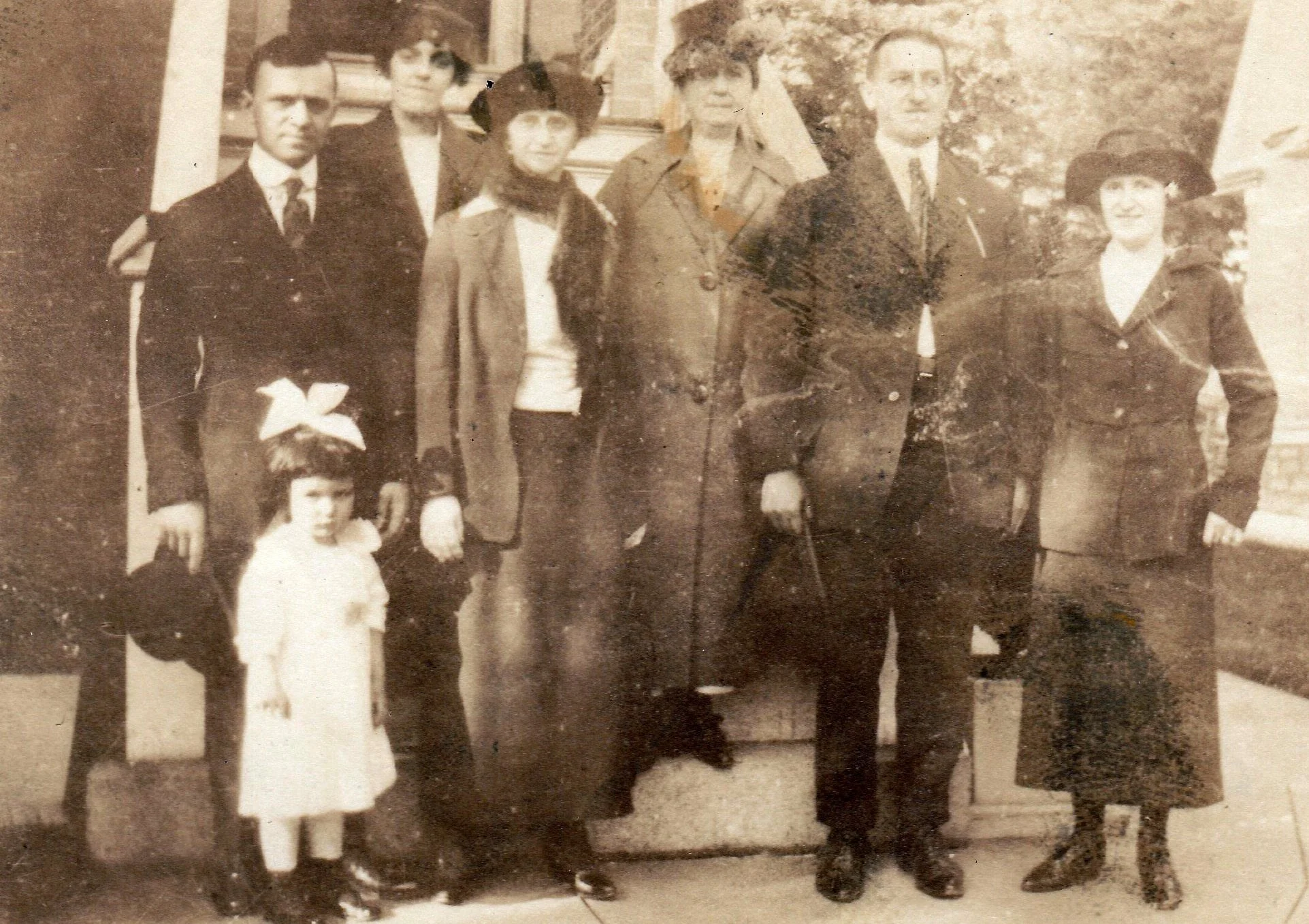 A vintage black and white photograph of a group of seven people, four adults and three children, standing outdoors in front of a house or porch. The adults are dressed in formal or period-appropriate clothing, including coats, hats, and skirts, while the children are in dressy outfits. The people are posing for the photo, with some smiling slightly.