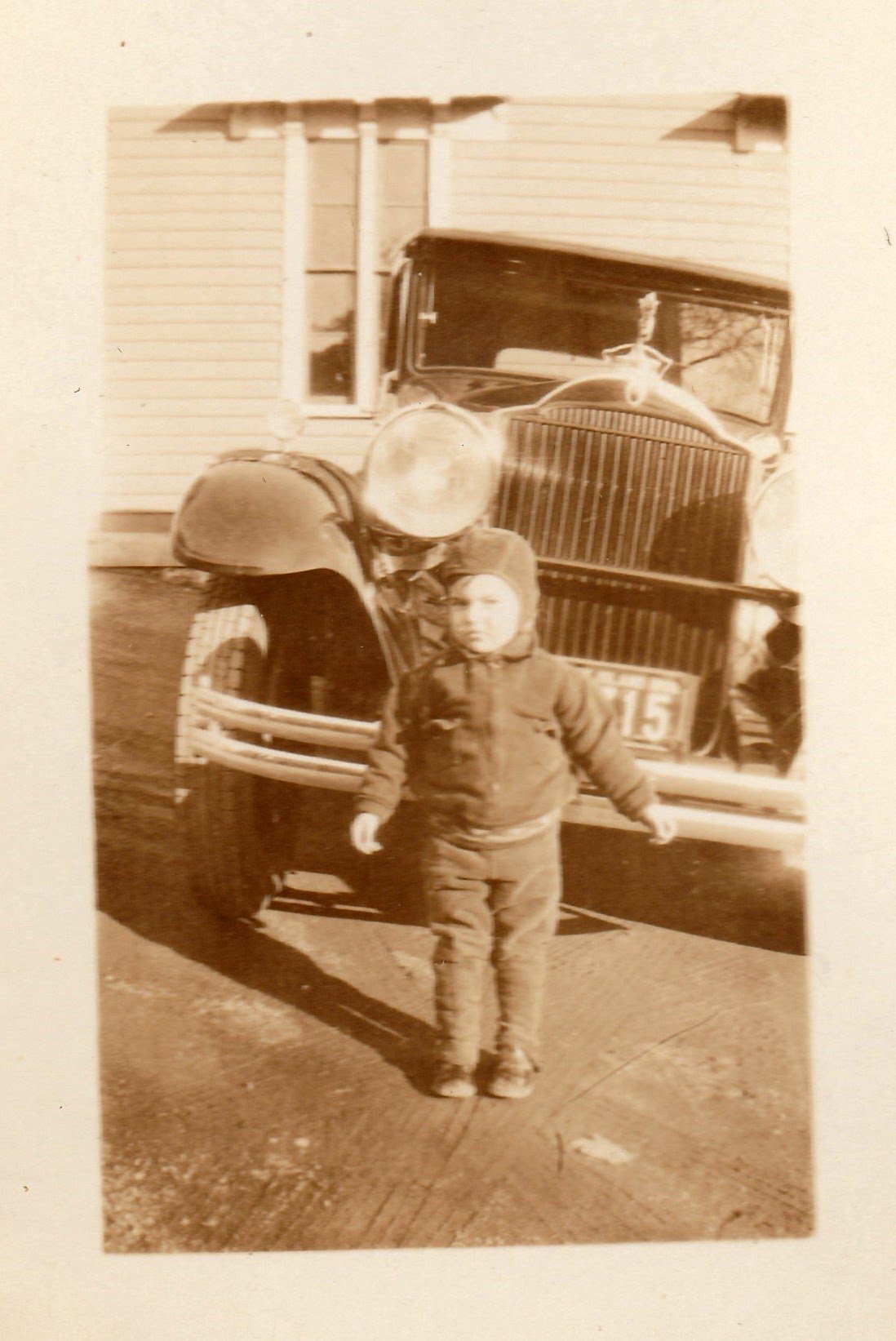 A young boy standing in front of a vintage fire truck with a large grille and round headlights, parked in front of a house with siding and a window.