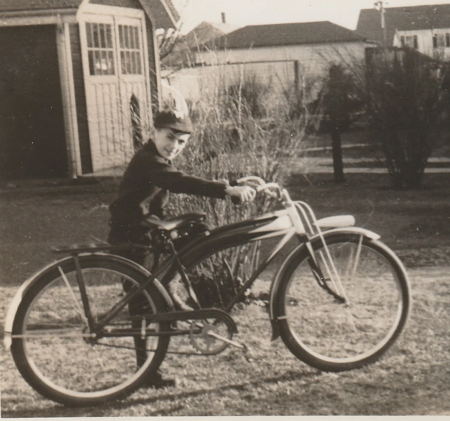 A young boy is sitting on a bicycle in a yard with grass, bushes, and houses in the background. The photo appears to be black and white.