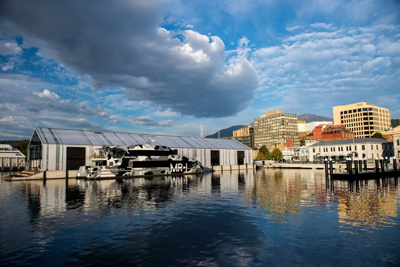 View of a waterfront city with a large warehouse-style building, a boat covered in a black and white camouflage pattern, and a skyline with various buildings against a partly cloudy sky.