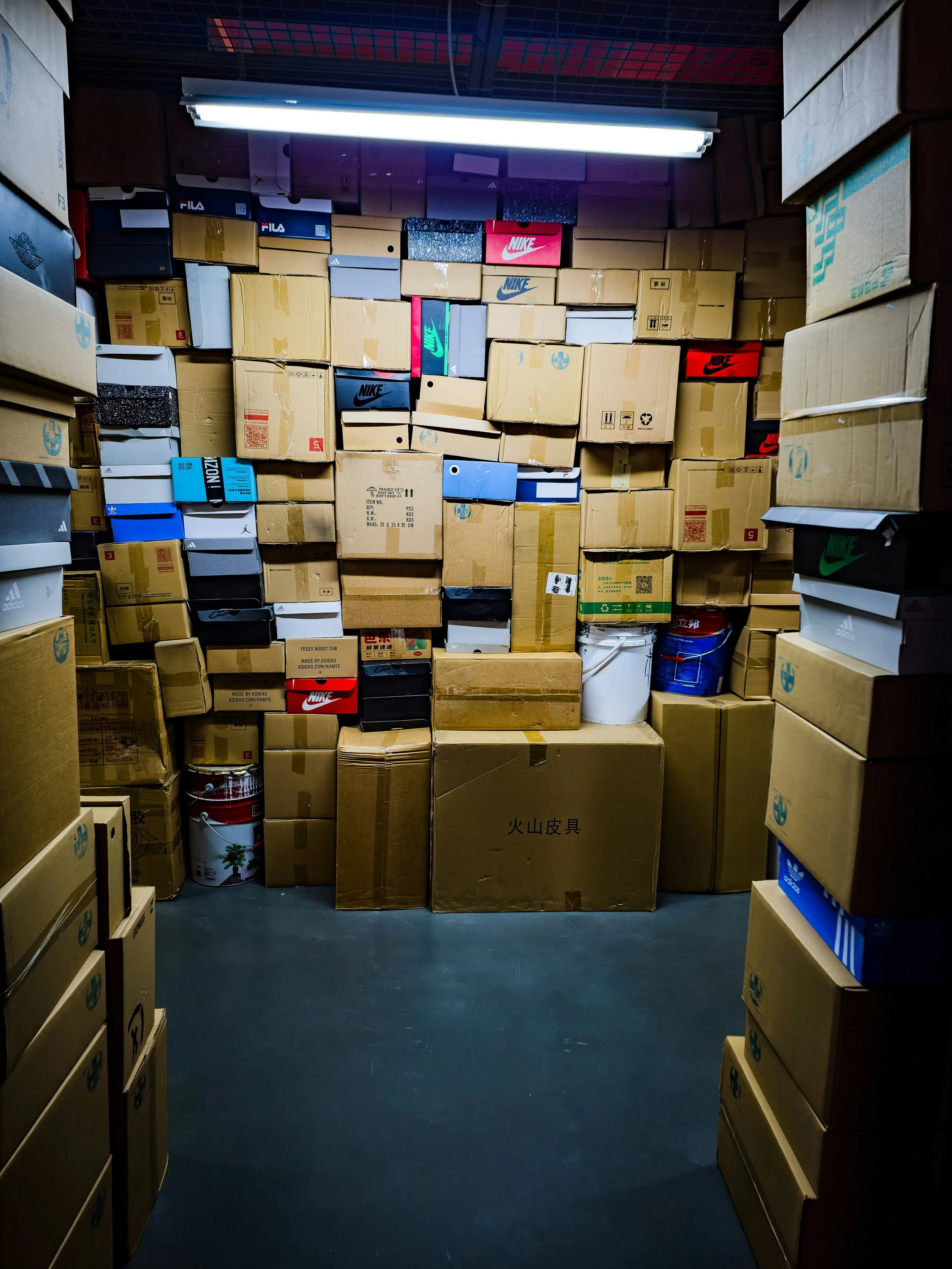 A storage room filled with stacked cardboard boxes of various sizes, brands, and colors, including Nike and Adidas, with a fluorescent light fixture on the ceiling illuminating the area.