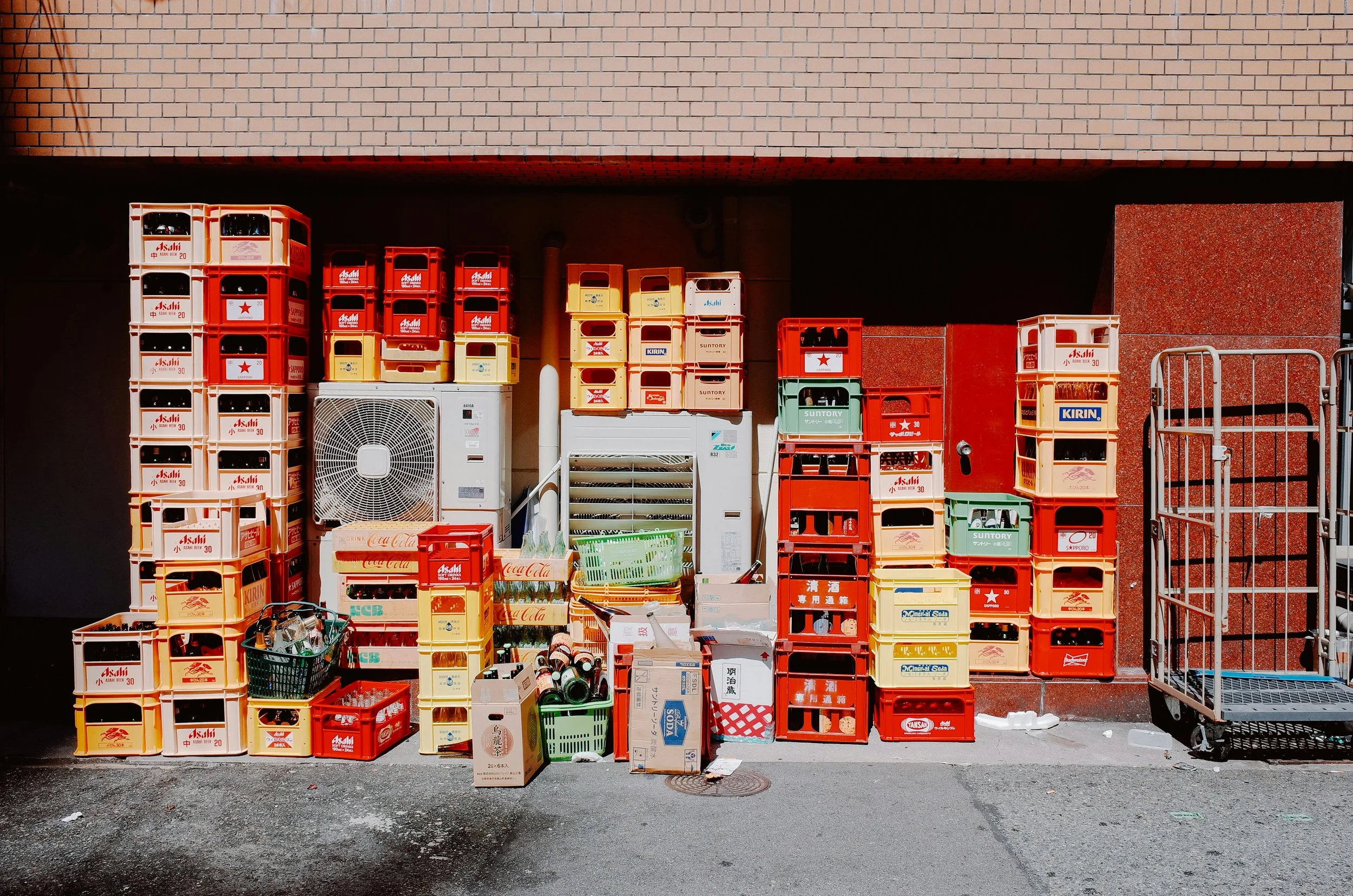 A stack of plastic crates and boxes, some with beverage logos, and a small shopping cart filled with bottles and cans, set against a brick wall and a sidewalk.