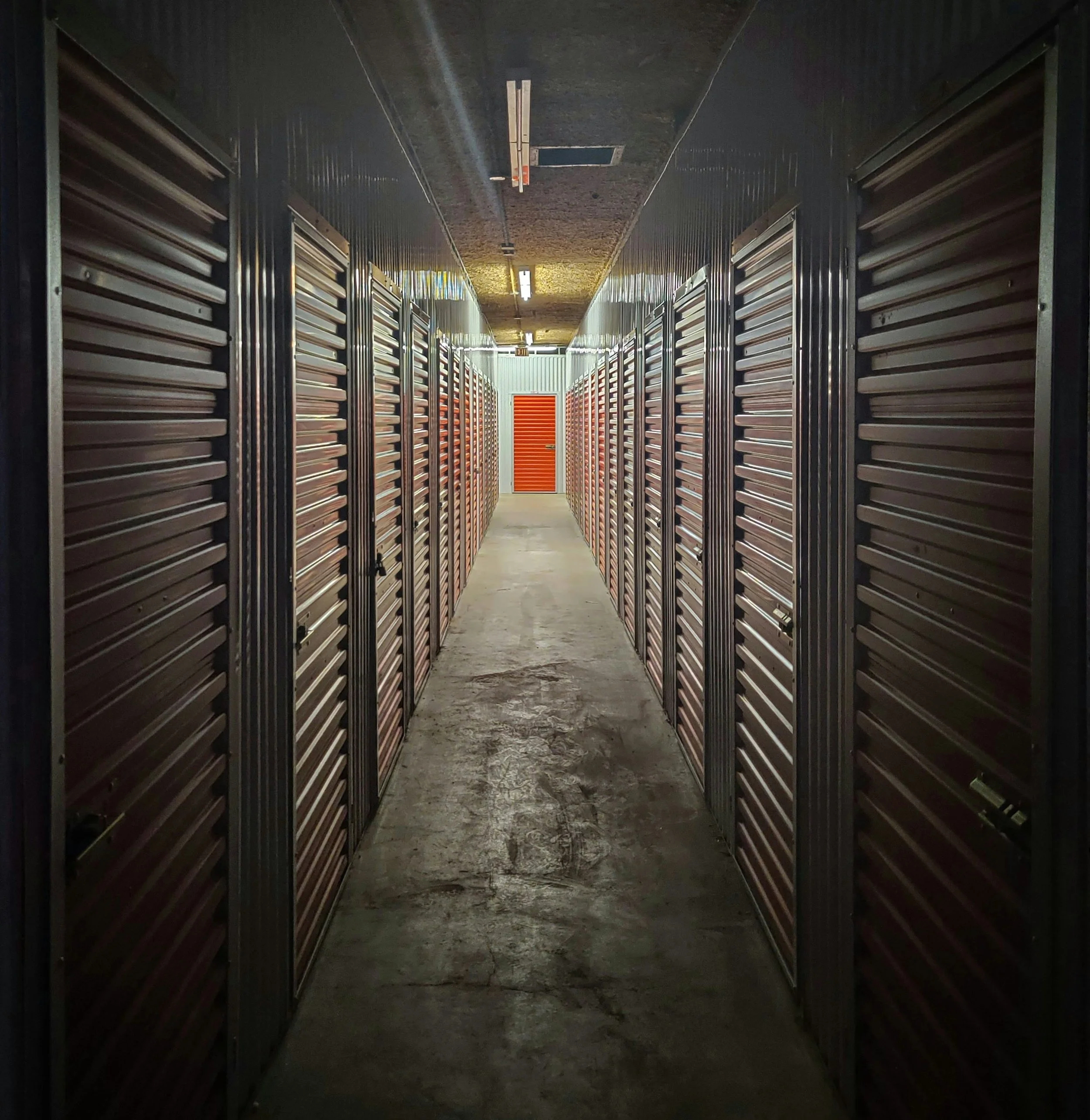 A hallway of storage units with orange doors, flanked by metal rolling doors on both sides, with a concrete floor and a ceiling with fluorescent lights.