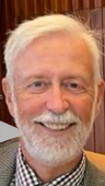 Close-up of a smiling older man with white hair and a beard, wearing a checkered shirt and a dark blazer.