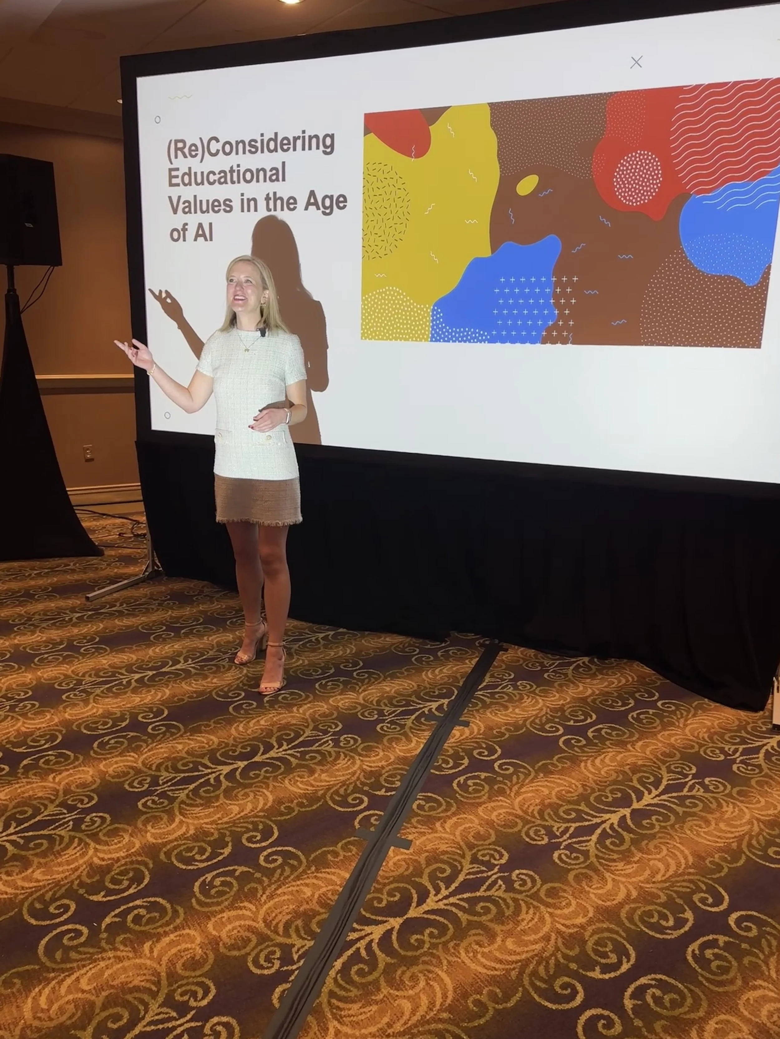 A woman, Amy Schumacher-Rutherford, standing and speaking at a conference, gesturing with her right hand, with a large presentation slide behind her displaying the title '(Re)Considering Educational Values in the Age of AI' and colorful abstract art.