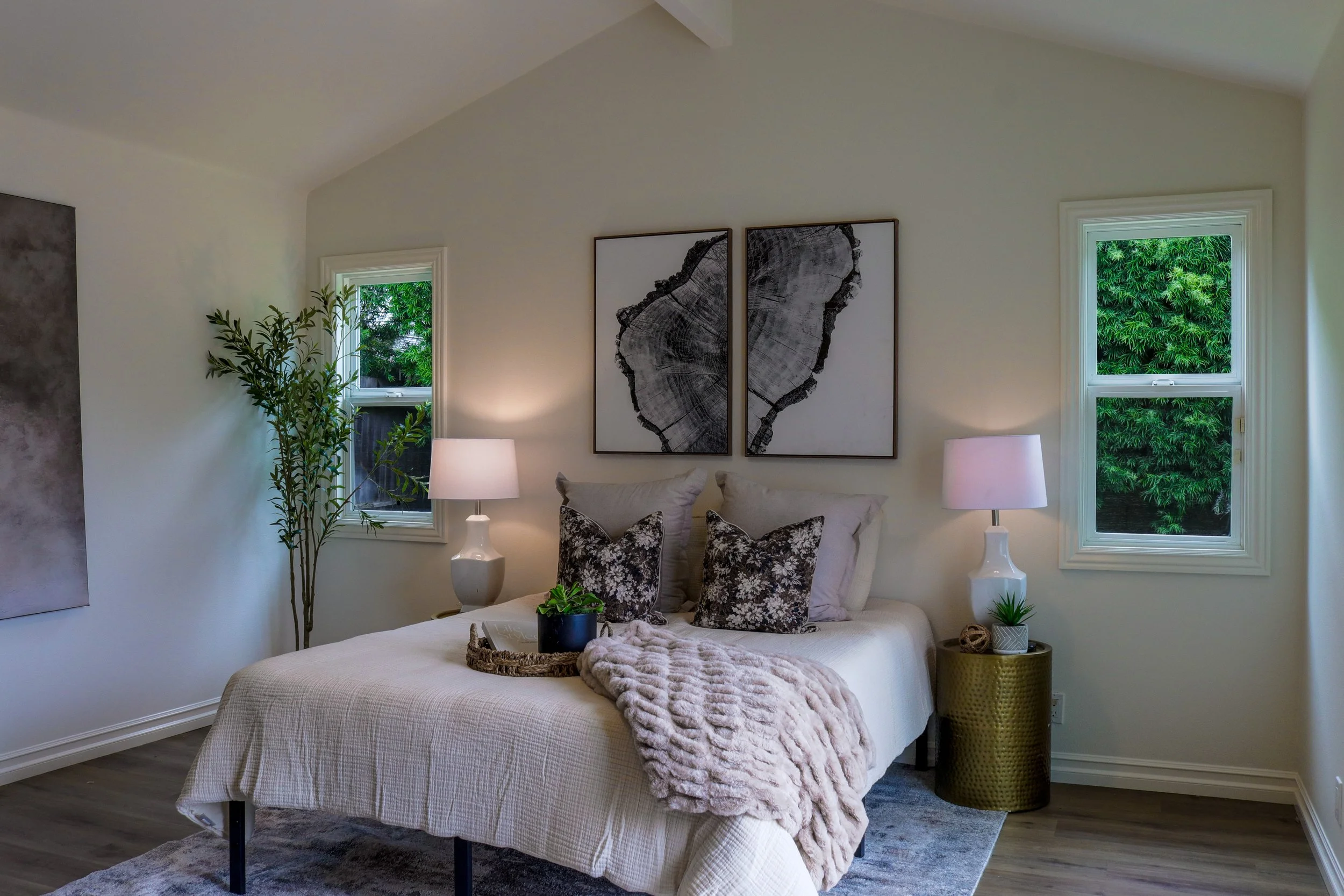 A cozy bedroom with white walls and two windows showing green foliage outside. The bed has beige bedding, patterned pillows, and a plush pink throw blanket. Two white lamps and decorative plants sit on nightstands beside the bed, with two large black