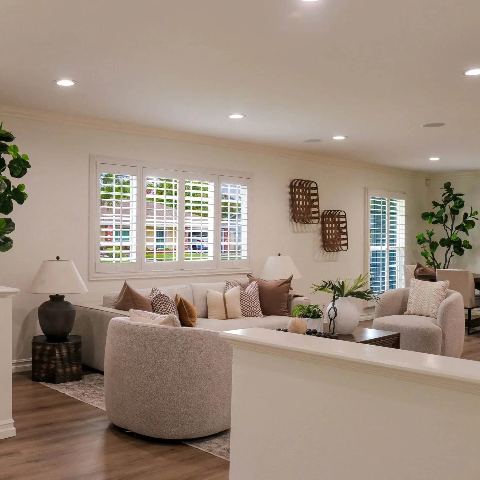 A living room with white walls, large windows with white shutters, a beige sofa with multiple pillows, and a white armchair. There is a wooden side table with a lamp, and decorative items including plants and wall art on the wall. Recessed lighting illuminates the room.