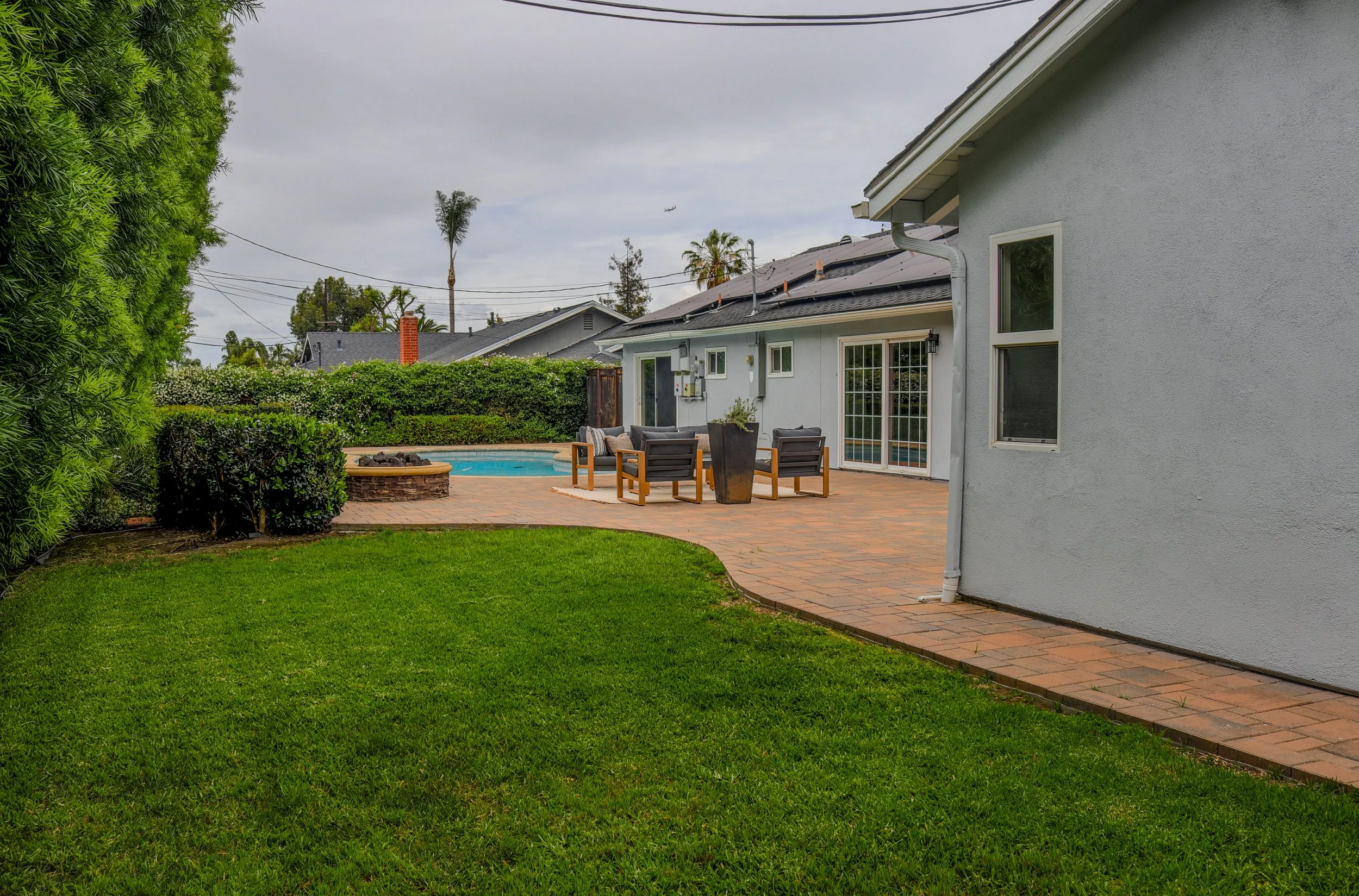 A backyard with a grassy lawn, a brick patio, an outdoor seating area with chairs and a tall planter, a swimming pool, lush green bushes, a gray house with a sliding door, and neighboring houses with palm trees in the background under an overcast sky