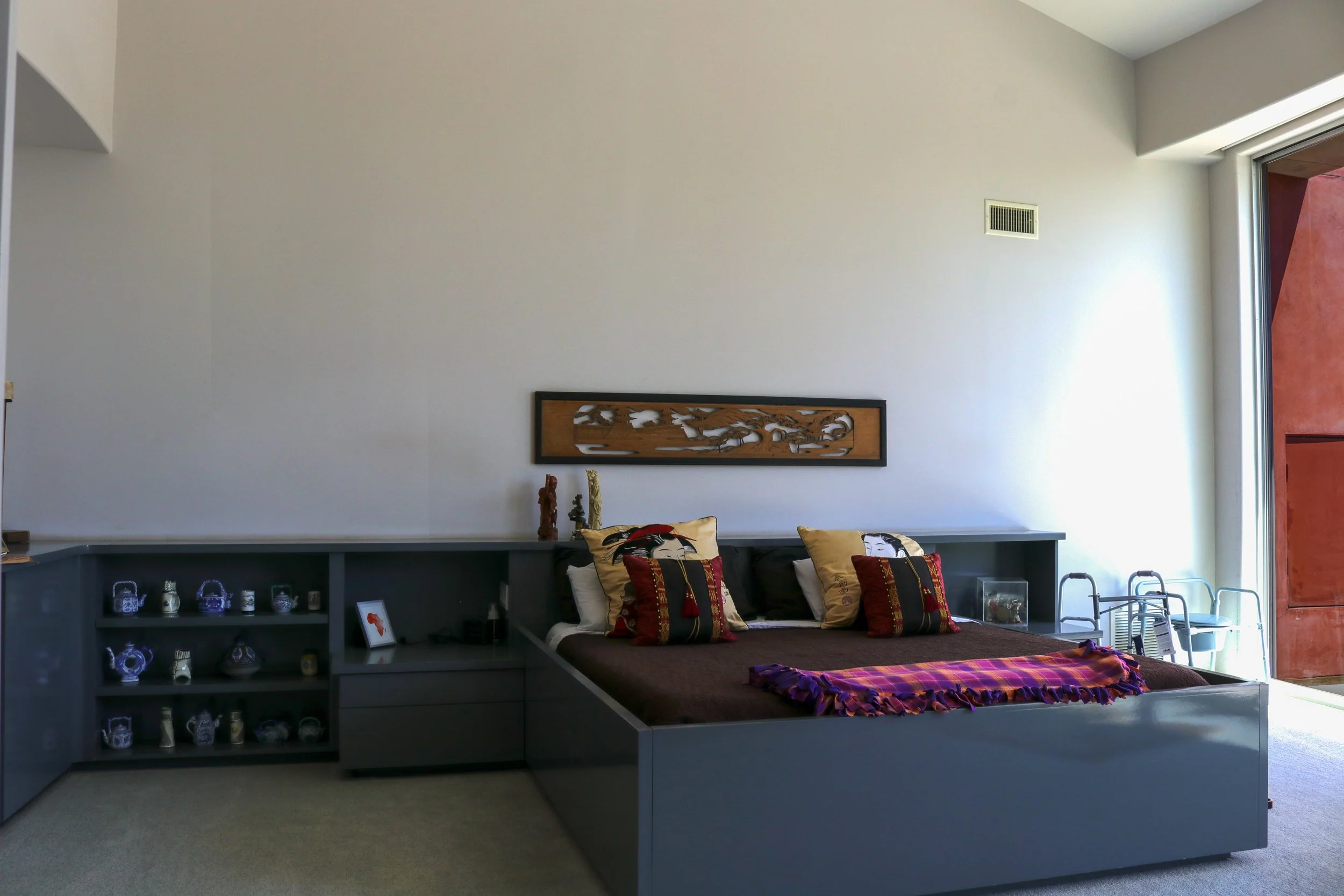 Modern living room with a gray platform bed, colorful pillows, a decorative wooden wall hanging, and a large window letting in natural light.