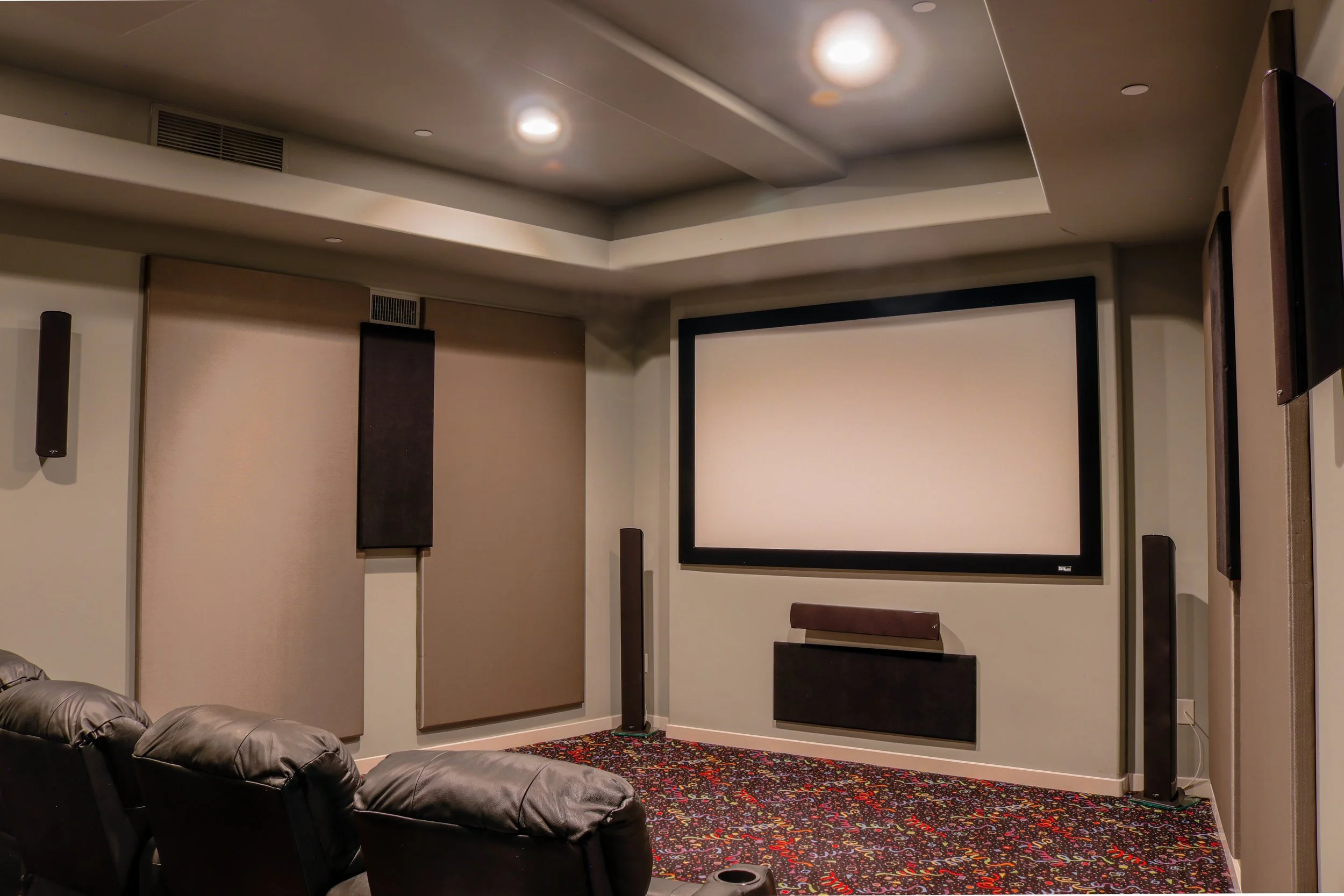 A home theater room featuring a large projection screen, multiple black speakers mounted on beige walls, carpeted floor with colorful specks, leather chairs, and a ceiling with built-in lighting.