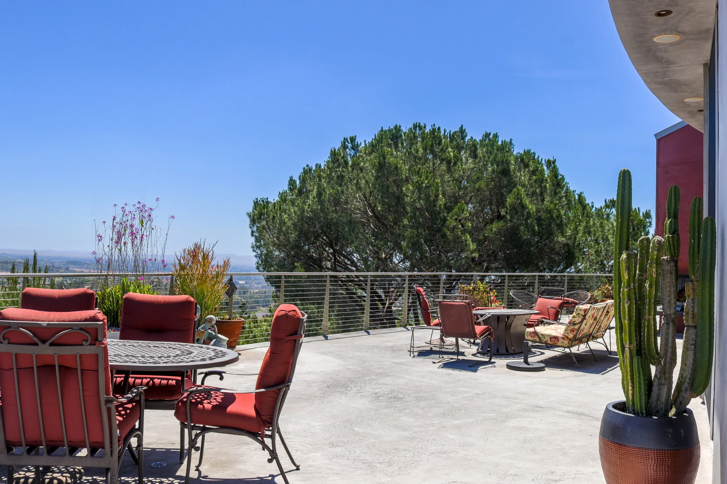 Outdoor patio with red cushioned chairs and tables, large potted cactus, plants, and a view of trees and city in the distance under a clear blue sky.