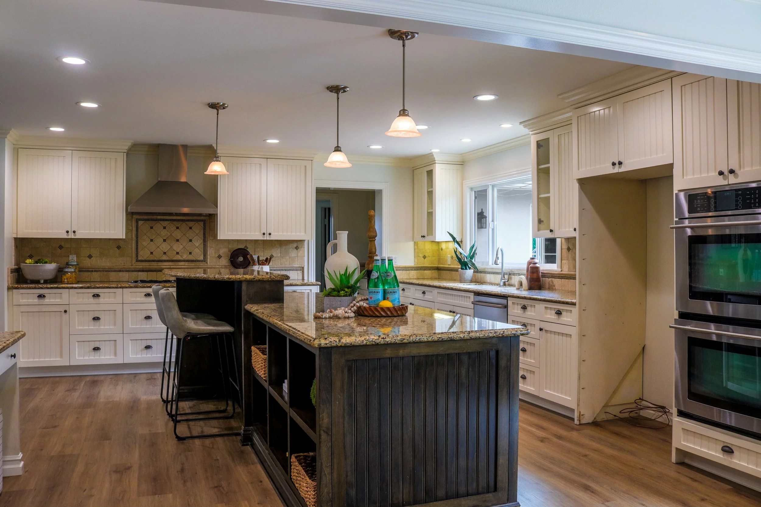A spacious kitchen with white cabinetry, a central island with a granite countertop, and stainless steel appliances. There are pendant lights hanging from the ceiling, and various decorative items and plants on the countertops.