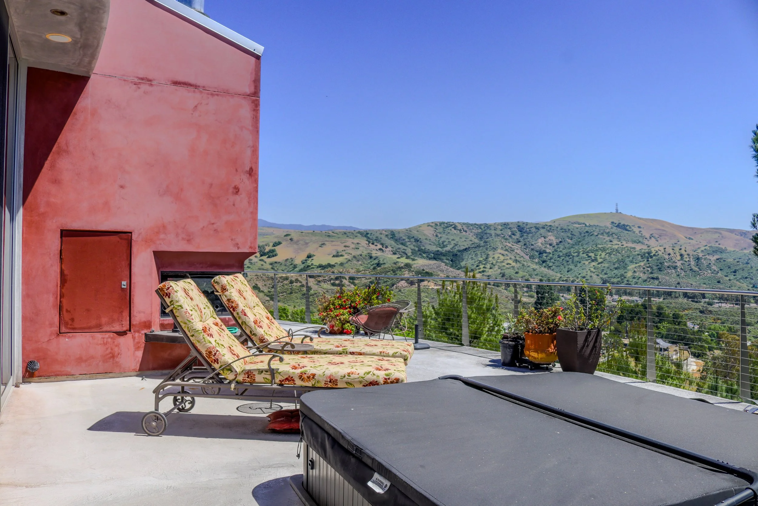 Apartment balcony with lounge chairs, potted plants, and mountain view with hills and clear blue sky.