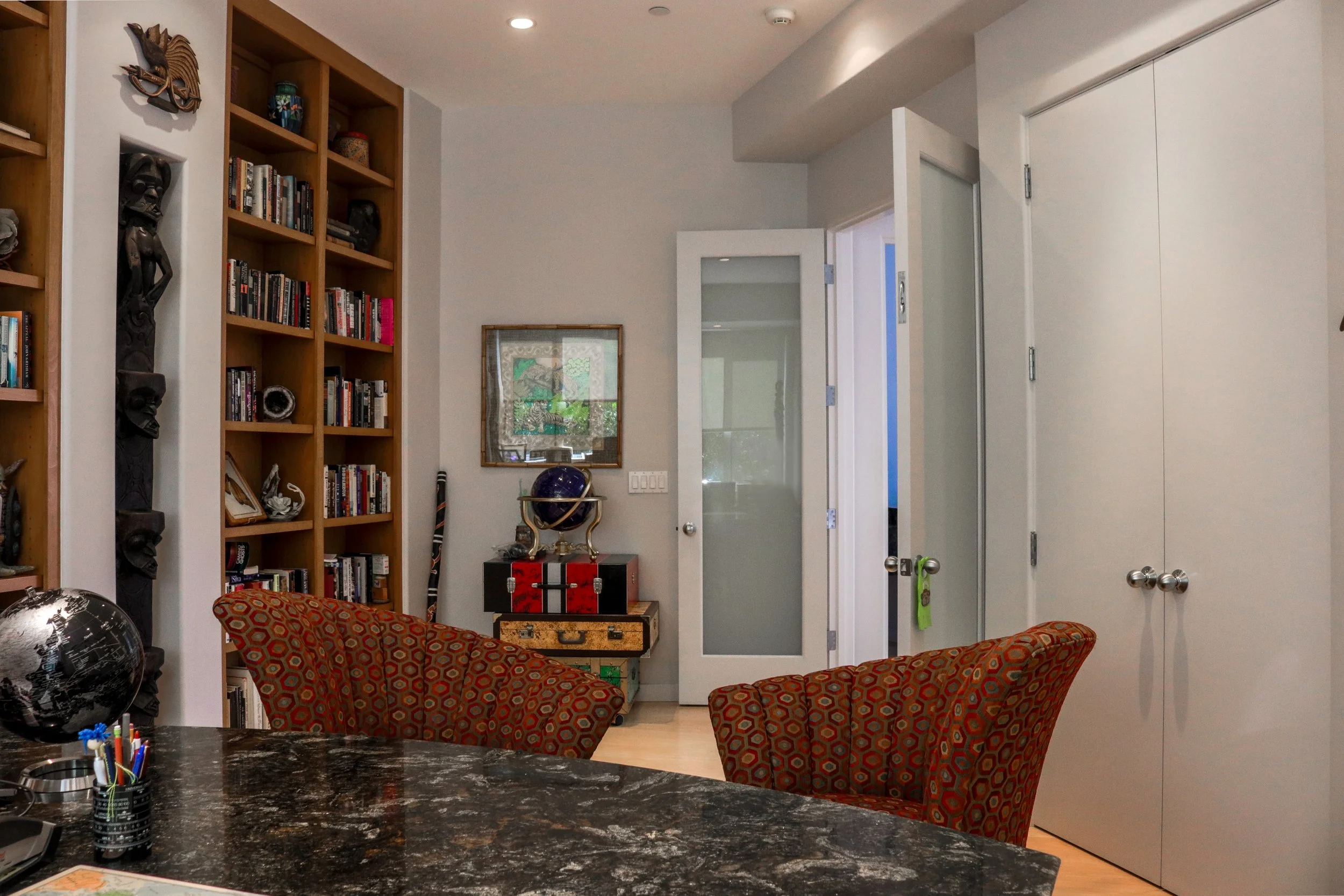 Interior of a modern house with white walls and furniture. There is a bookshelf with books and decorations, a small table with a globe, and two patterned armchairs. An open door with frosted glass leads to another room.