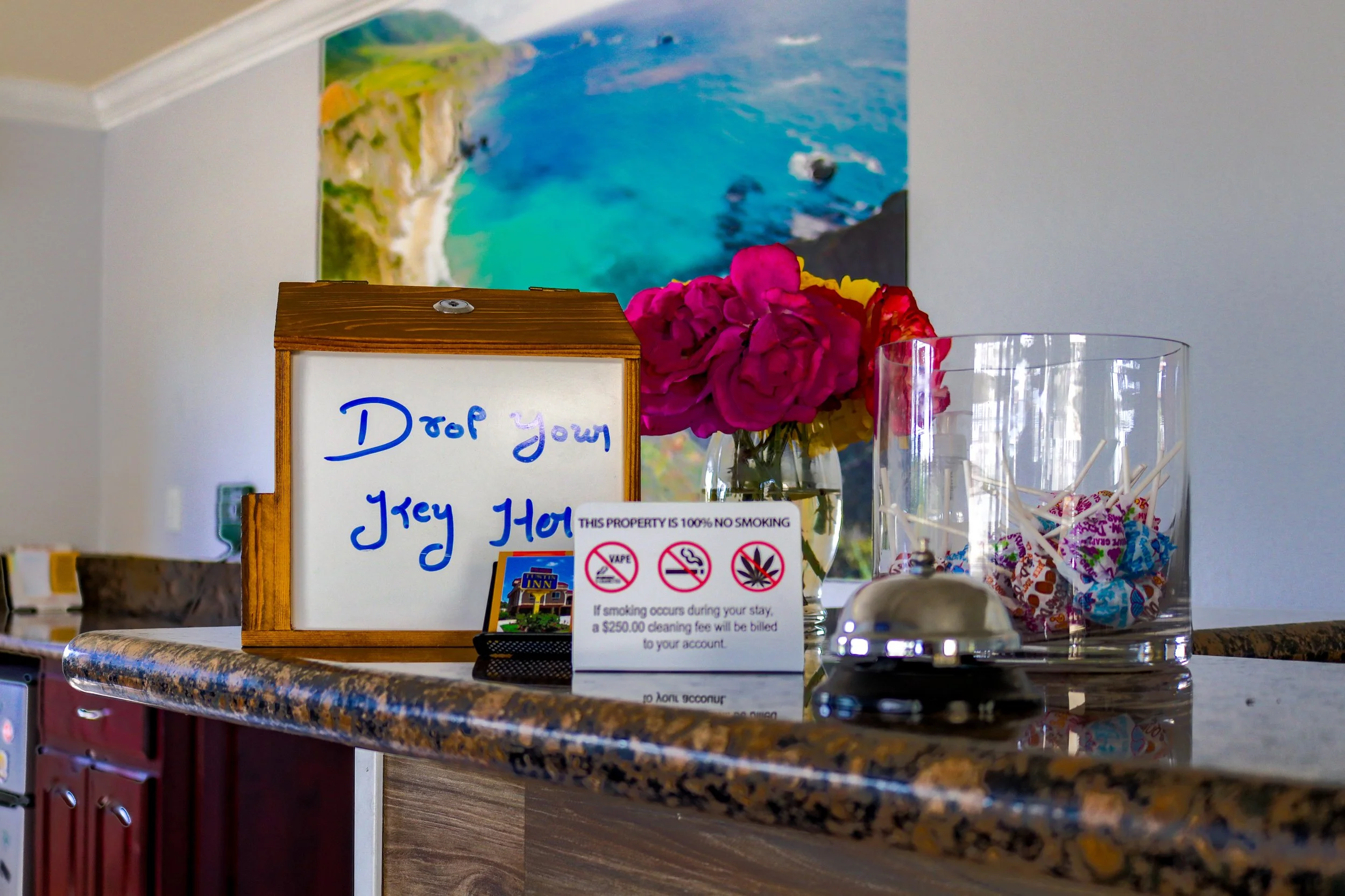 Decorative counter with pink and yellow flowers, glass jar, and a whiteboard with writing and signs, setting in a kitchen with a colorful landscape picture on the wall.