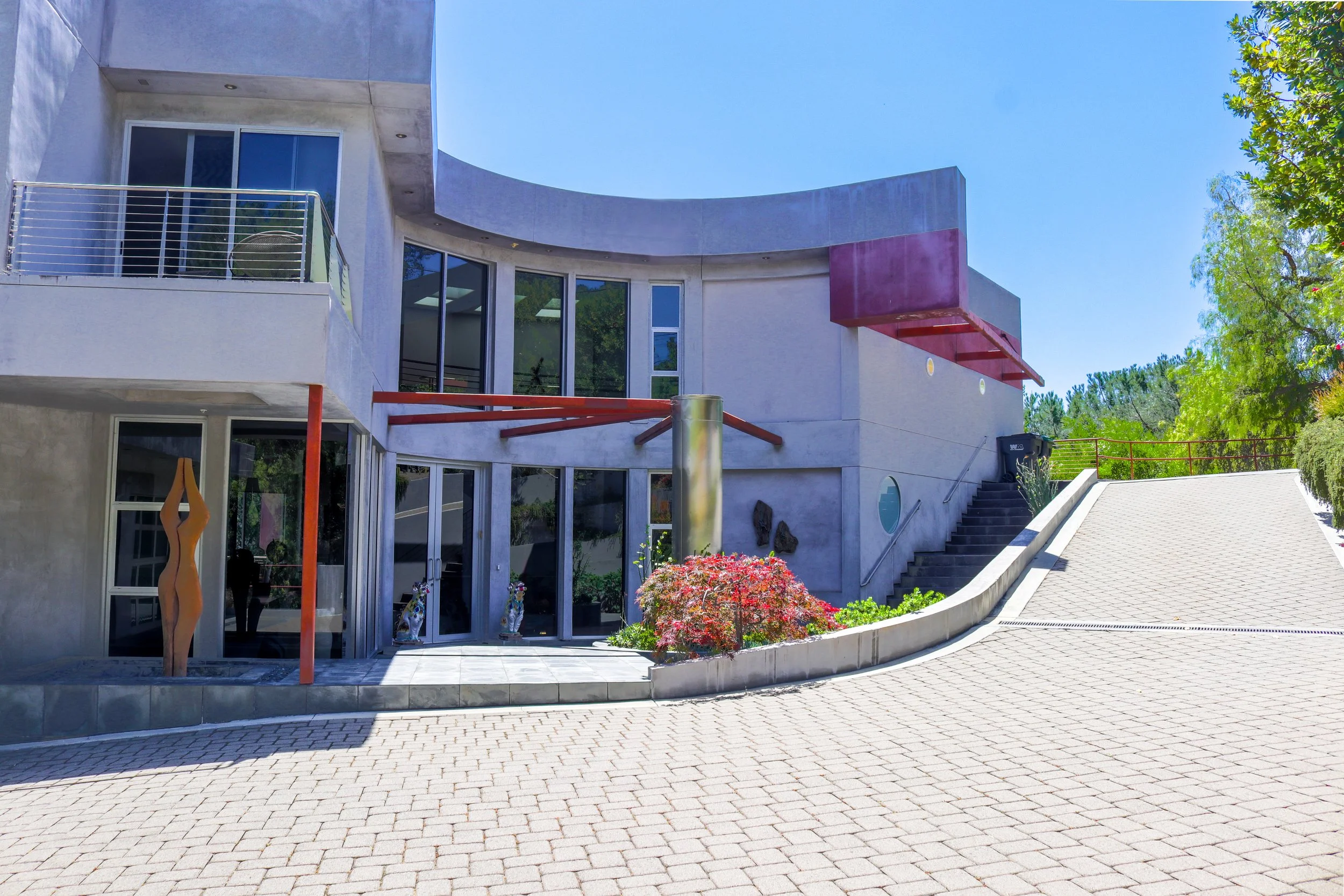 Modern multi-story house with white exterior walls, large windows, and a curved upper balcony. There are trees and plants around the house, and a paved driveway in front.