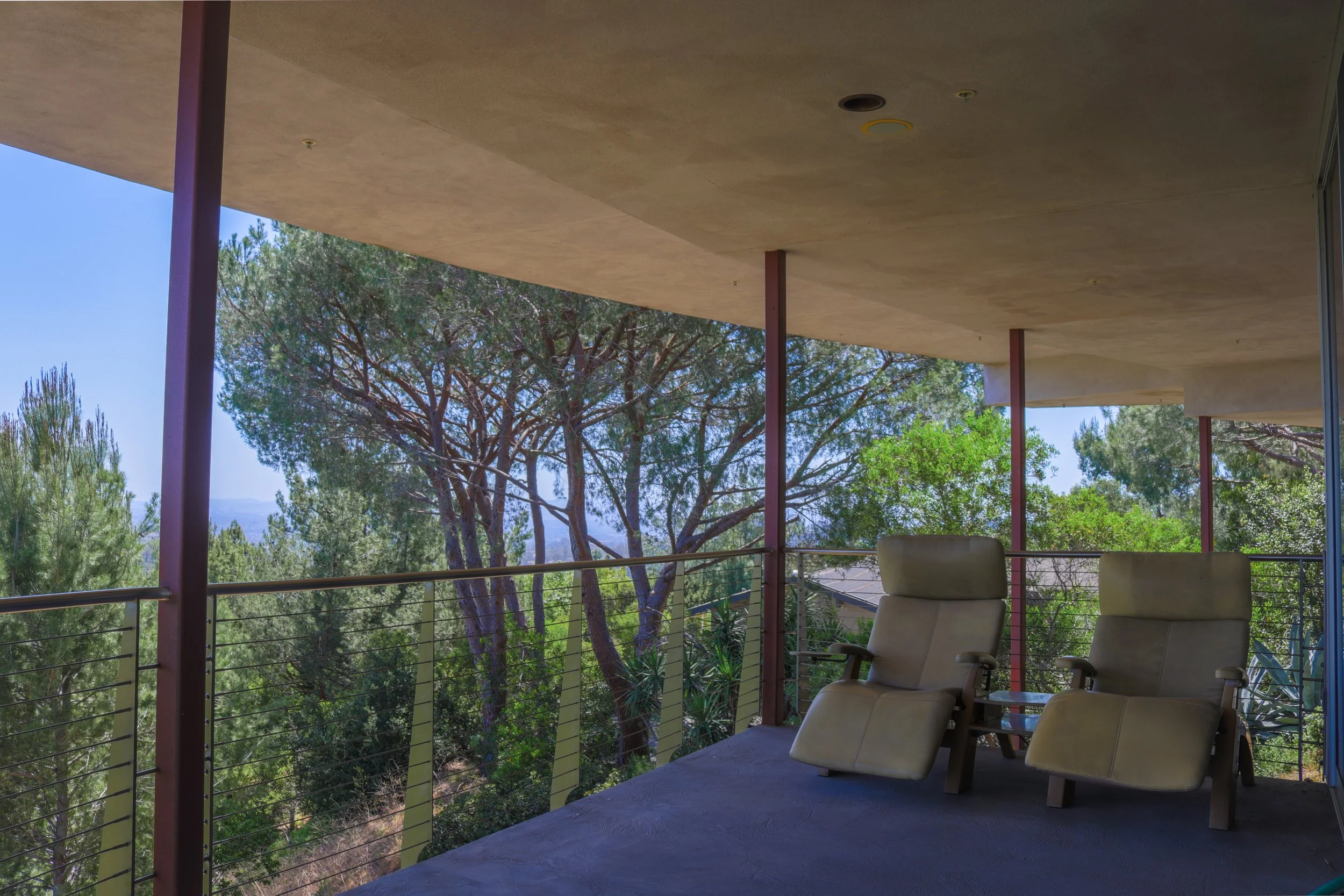 A covered balcony with beige chairs, metal railing, and a view of trees with green foliage and a clear blue sky.