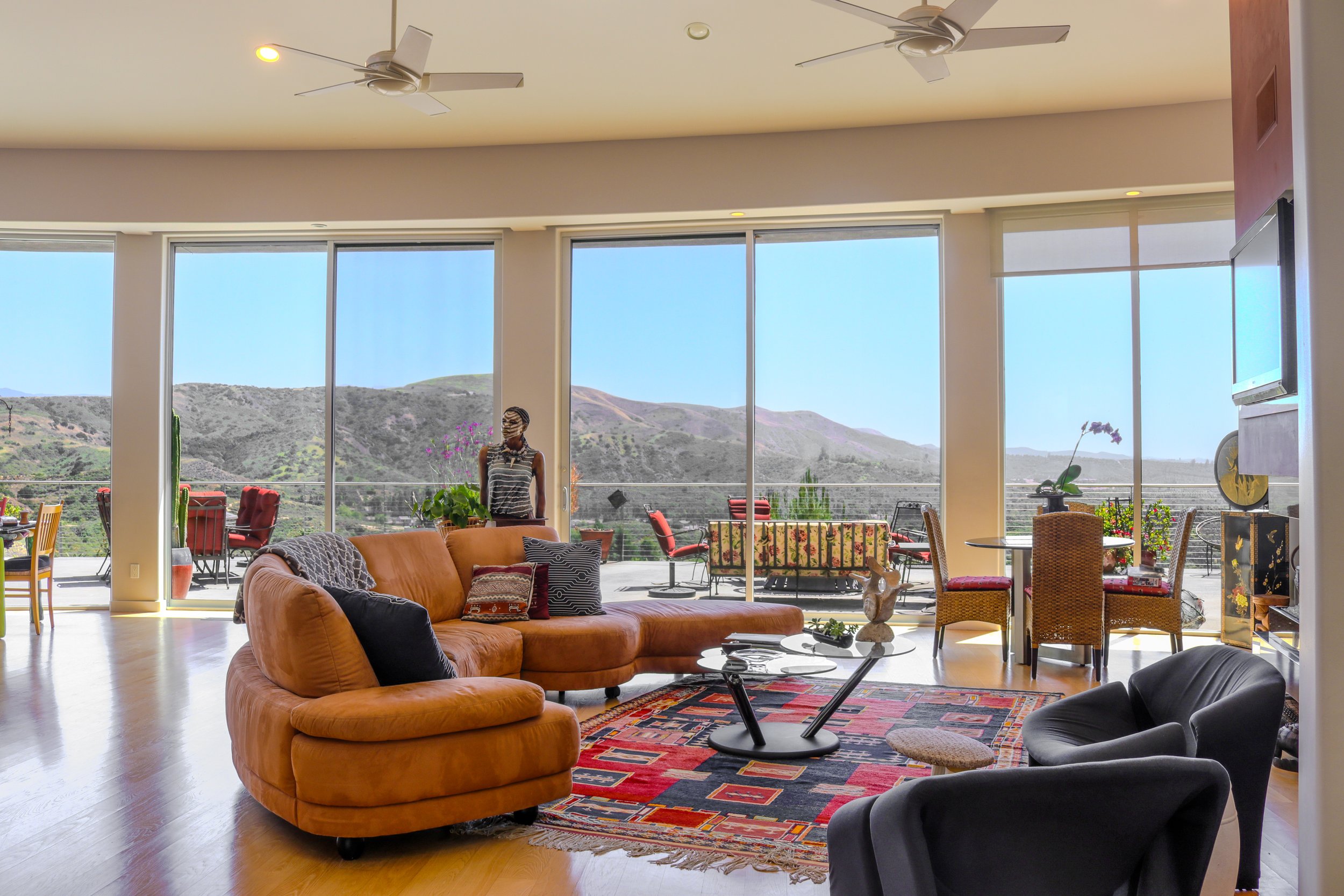 A spacious living room with large glass windows overlooking mountains, furnished with a mix of sofas, chairs, a coffee table, and decorative plants, with a person standing near the windows.