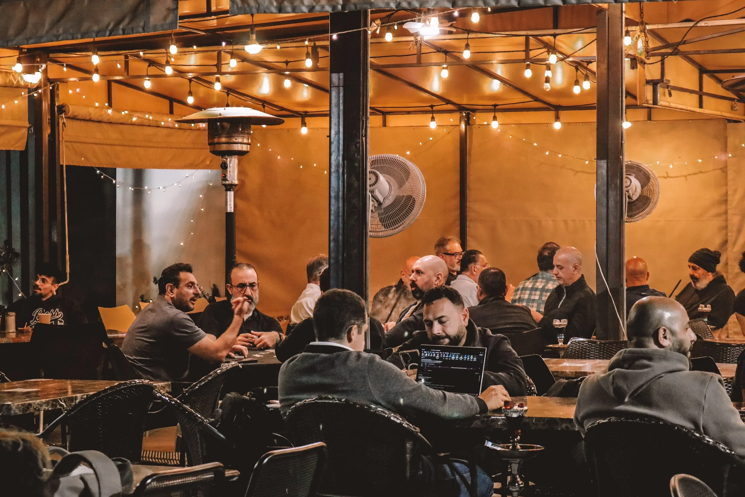 Indoor social gathering with several people sitting at tables, some using laptops, under warm yellow lighting with string lights and fans in the background.