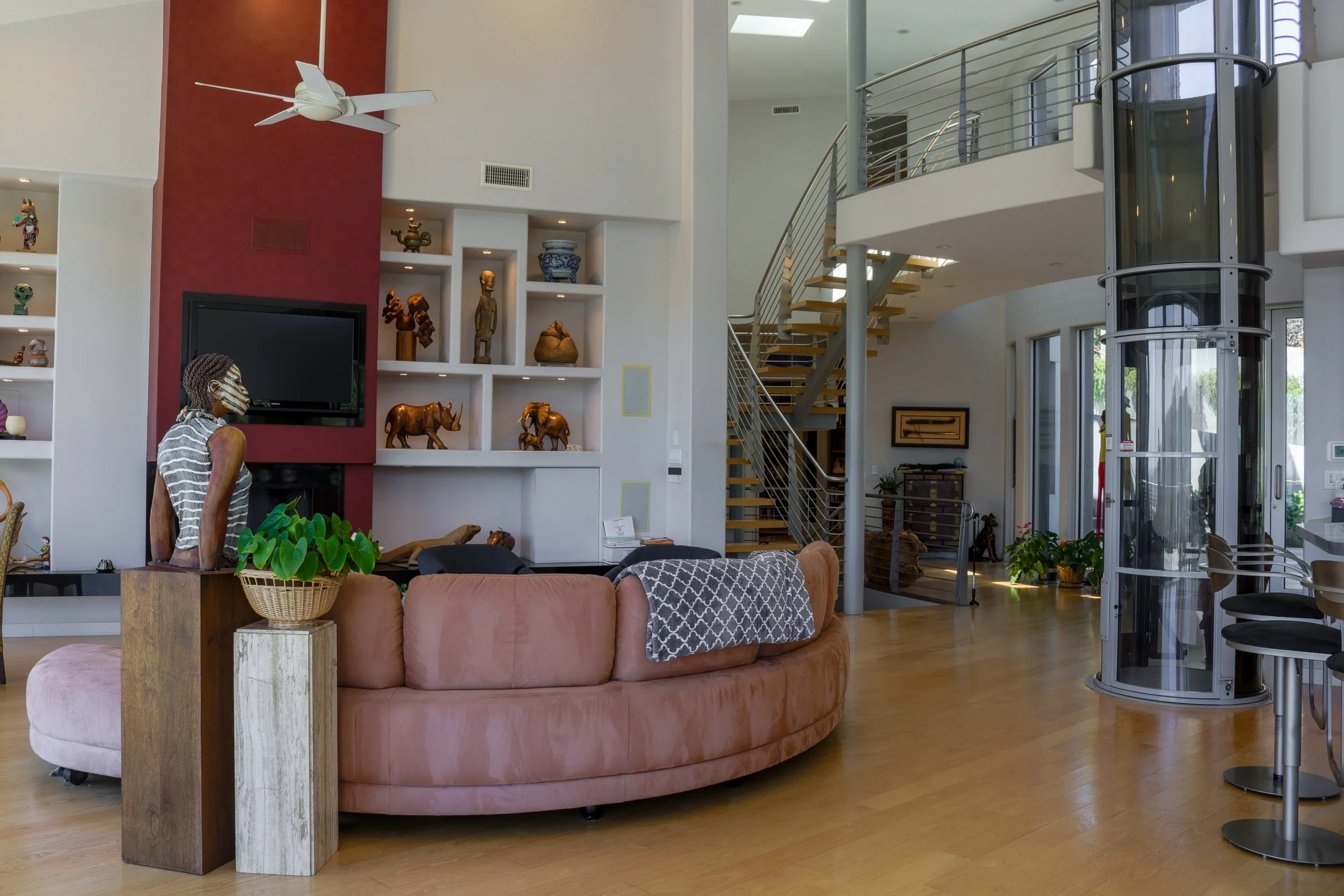 Living room with a pink sofa, wooden side table, eclectic sculpture, bookshelves with decorative items, revolving elevator, staircase, and large windows, in a modern house.