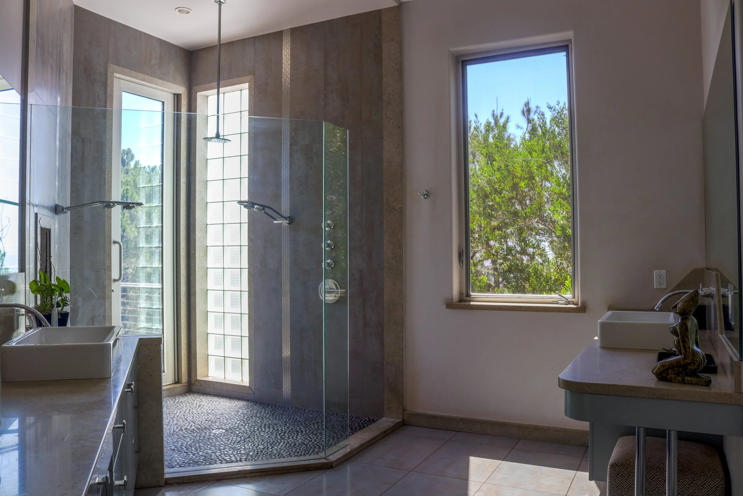 Modern bathroom with large window, walk-in shower with glass enclosure, white sink, and countertop with decorative items, and a small plant.